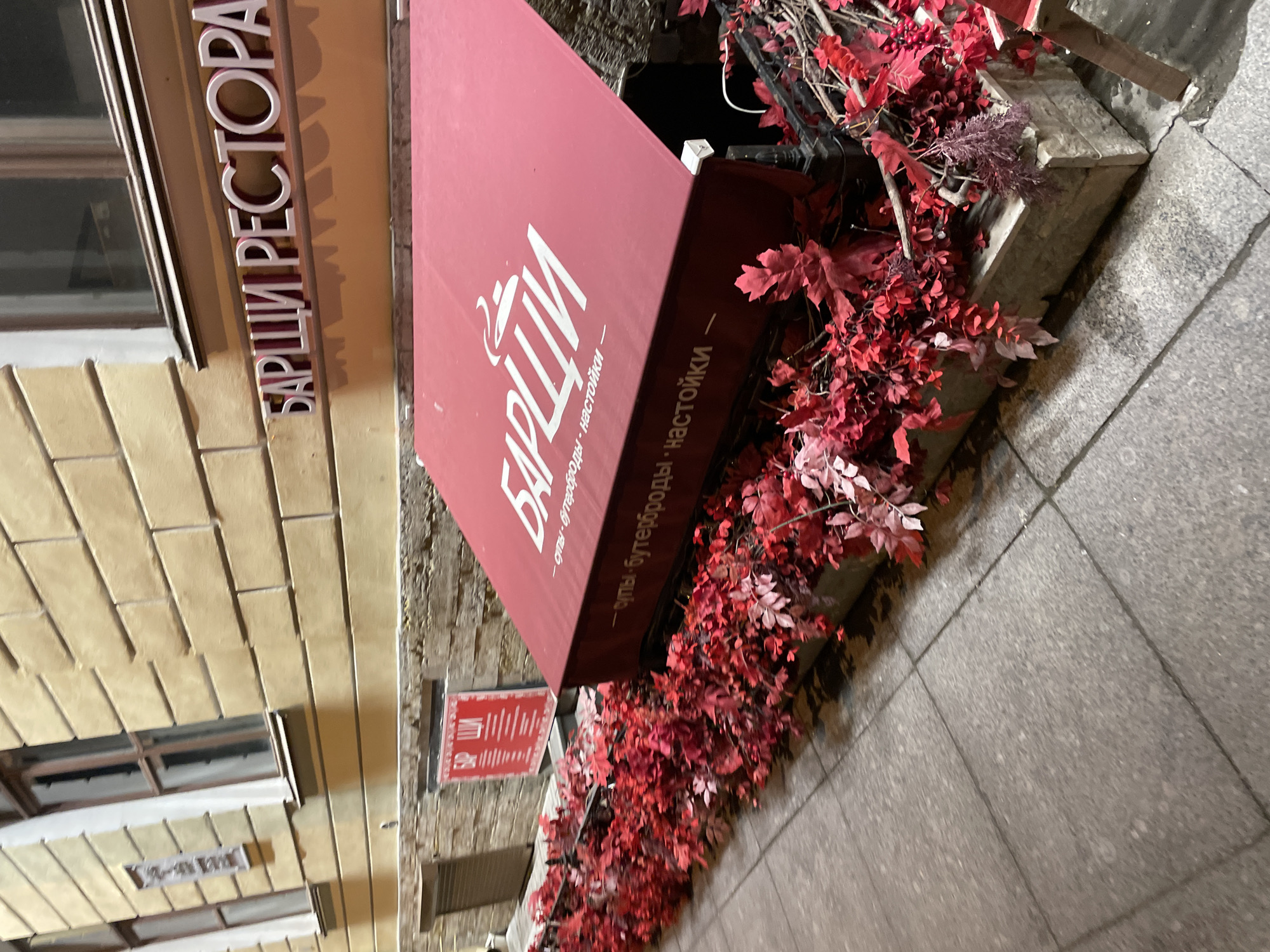 Here is a description of the image:
The image shows an exterior view of a restaurant named "БАР ШИ РЕСТОРАН" which translates to "BAR SHI RESTAURANT". A red awning above the entrance of the restaurant reads "БАР ШИ - супы • бутерброды • настойки" which translates to "BAR SHI - Soups • Sandwiches • Tinctures". The exterior wall of the restaurant is made of yellow bricks. The building appears to be a historical structure, typical of Saint Petersburg, Russia. The entrance to the restaurant is adorned with red foliage, possibly autumnal decor, cascading down the front of the building onto the pavement. The location is likely within the city of Saint Petersburg, Russia due to the Cyrillic signage and architectural style. There are no people visible in the shot. The lighting suggests the photo was taken in the evening or during twilight.