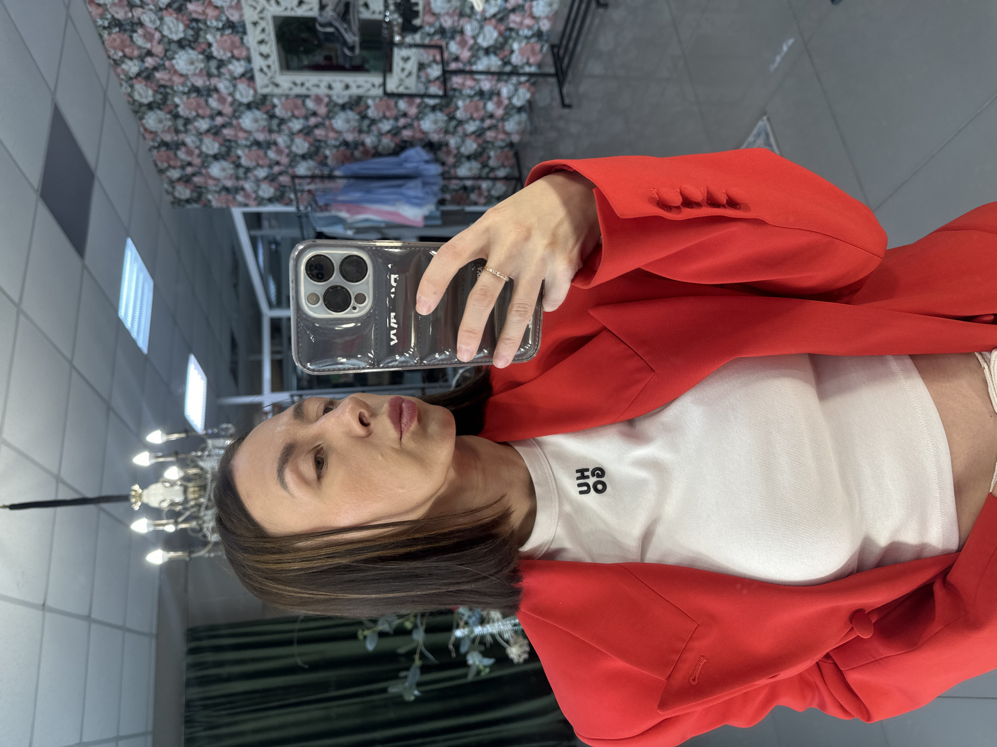 A woman with dark hair, wearing a red blazer over a white t-shirt, takes a selfie in a store. The t-shirt has a small black logo that appears to say "UH OO." She is wearing a ring on her right hand. The background shows a wall with floral wallpaper, clothing racks with various garments, and a chandelier. The floor is tiled. The lighting suggests an indoor setting, possibly a boutique. There is no explicit indication of the location being Votkinsk, Russia.