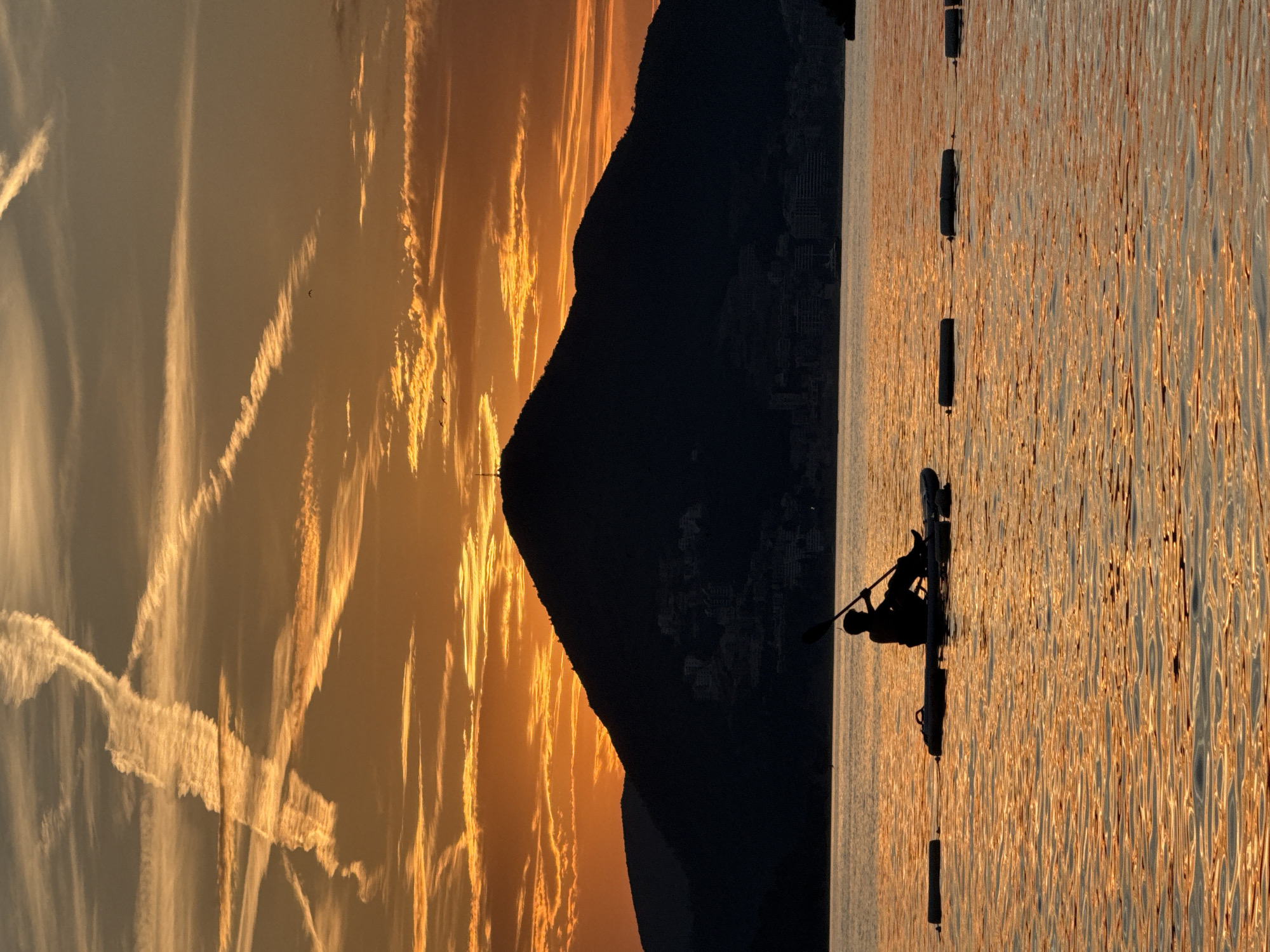 A lone kayaker, silhouetted against a vibrant orange sunset, paddles on the calm waters of Pržno, Montenegro. The sky is filled with dramatic, streaky clouds, some illuminated by the setting sun, and airplane contrails crisscross the upper atmosphere. In the background, a dark, mountainous silhouette looms, with scattered lights indicating a town nestled on its slopes. The water reflects the fiery hues of the sky, creating a shimmering golden surface. A line of buoys is visible in the foreground, marking a section of the sea. The kayaker is seated on a paddleboard, holding a paddle, and appears to be calmly enjoying the serene twilight.