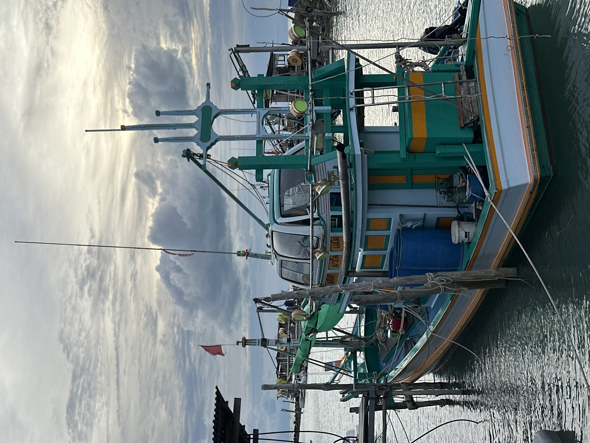 A fishing boat is docked in a harbor in the Ko Pha-ngan District of Thailand. The boat is green, white, and yellow, with an enclosed cabin and a mast. There are several lights mounted on the boat. In the background, other boats and structures can be seen. The sky is cloudy, and the water is calm.