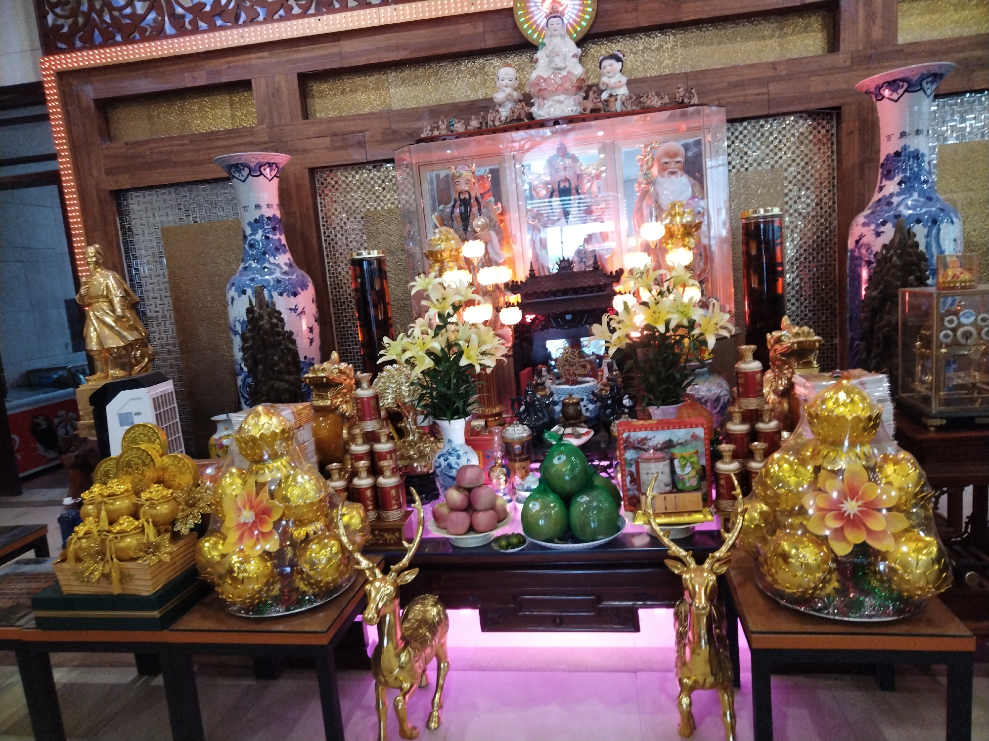 Captured indoors in Chu Văn An Ward, Vietnam, the image showcases a detailed Buddhist altar. Two tables flank the scene; the left table presents gold-colored artifacts, including pots filled with coins, while the right mirrors this with similar items, both protected by clear plastic wrap. Between them sits a central table laden with offerings, including fresh fruits like apples and pomelos arranged in bowls. Two gold-colored deer statuettes guard each side of this table. The backdrop of the altar features elaborately decorated vases and religious figures within a glass-fronted cabinet, all framed by dark wood and punctuated by subtle lighting. The overall environment suggests a place of reverence and cultural practice.