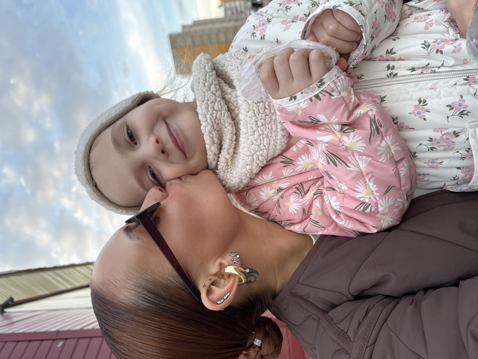A selfie shows a woman and a child in an outdoor setting. The woman, wearing brown sunglasses and a brown jacket, is holding the child, who is wearing a white hat, a white scarf, and a pink and white floral jacket. The woman is kissing the child on the cheek. The background features a building and a cloudy sky. The location is Chaikovsky, Russia.