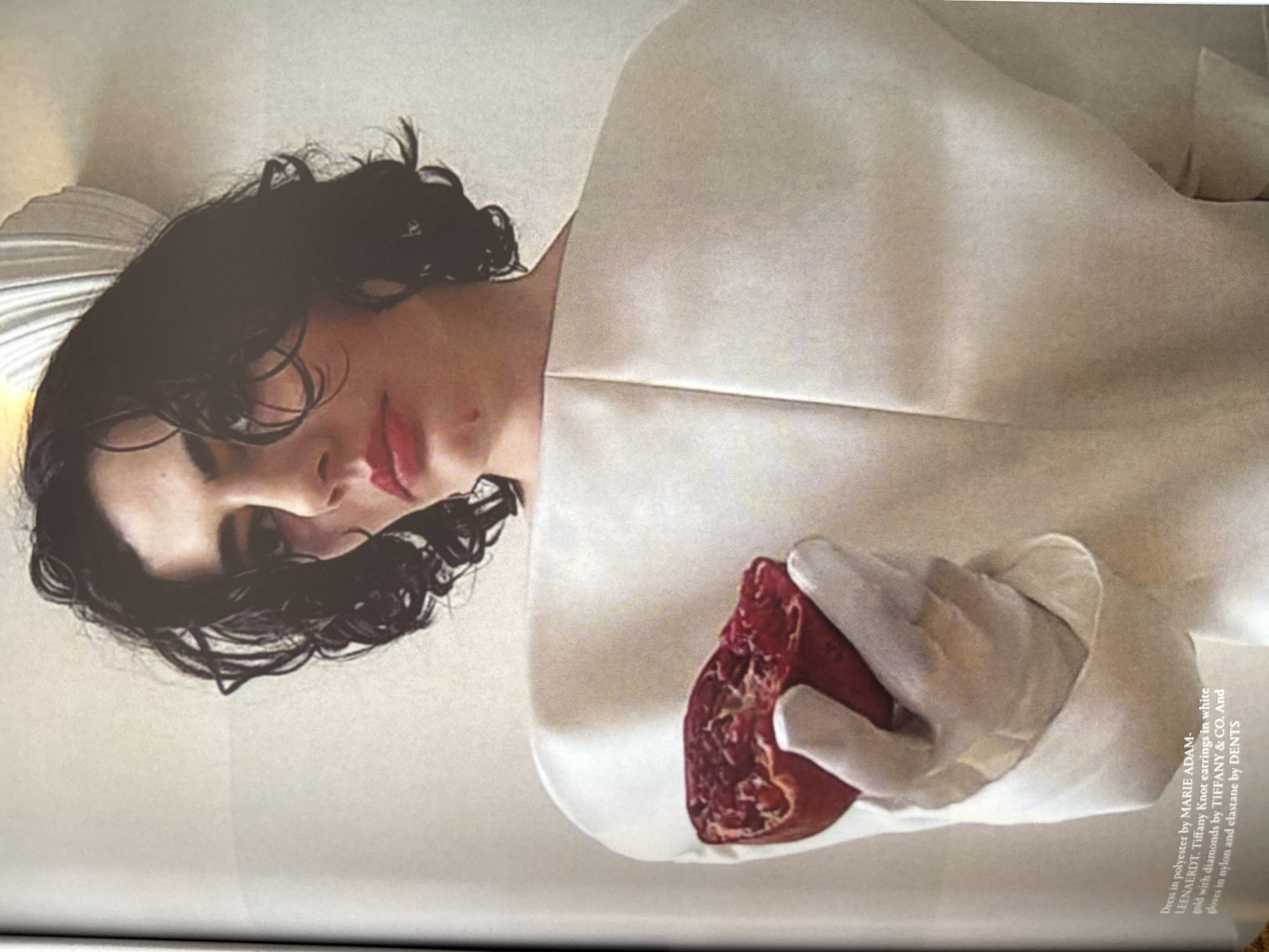 A person with dark, curly hair and light skin lies on a light-colored surface, wearing a white garment. The person holds a pomegranate in their gloved hand. The text indicates the clothing is by Marie Adam-Leenaerdt, earrings by Tiffany & Co., and gloves by DENTS. The scene does not appear to be located in London, United Kingdom. There is no readily apparent cityscape or landmark.