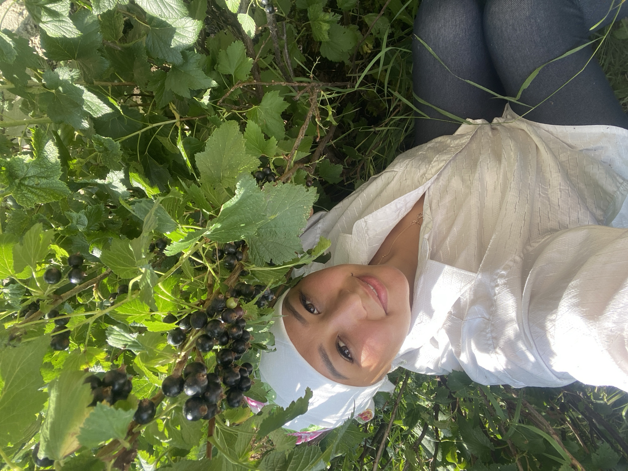 In the village of Maloarslangulovo, Russia, a woman is pictured lying down amidst a blackcurrant bush. She is wearing a white headscarf with floral accents, a white button-down shirt, and dark pants. The blackcurrant bush is laden with ripe, dark berries and green leaves. Sunlight filters through the foliage, casting dappled shadows on the scene. The woman is looking directly at the camera, smiling slightly. The overall setting appears to be a garden or natural outdoor space.