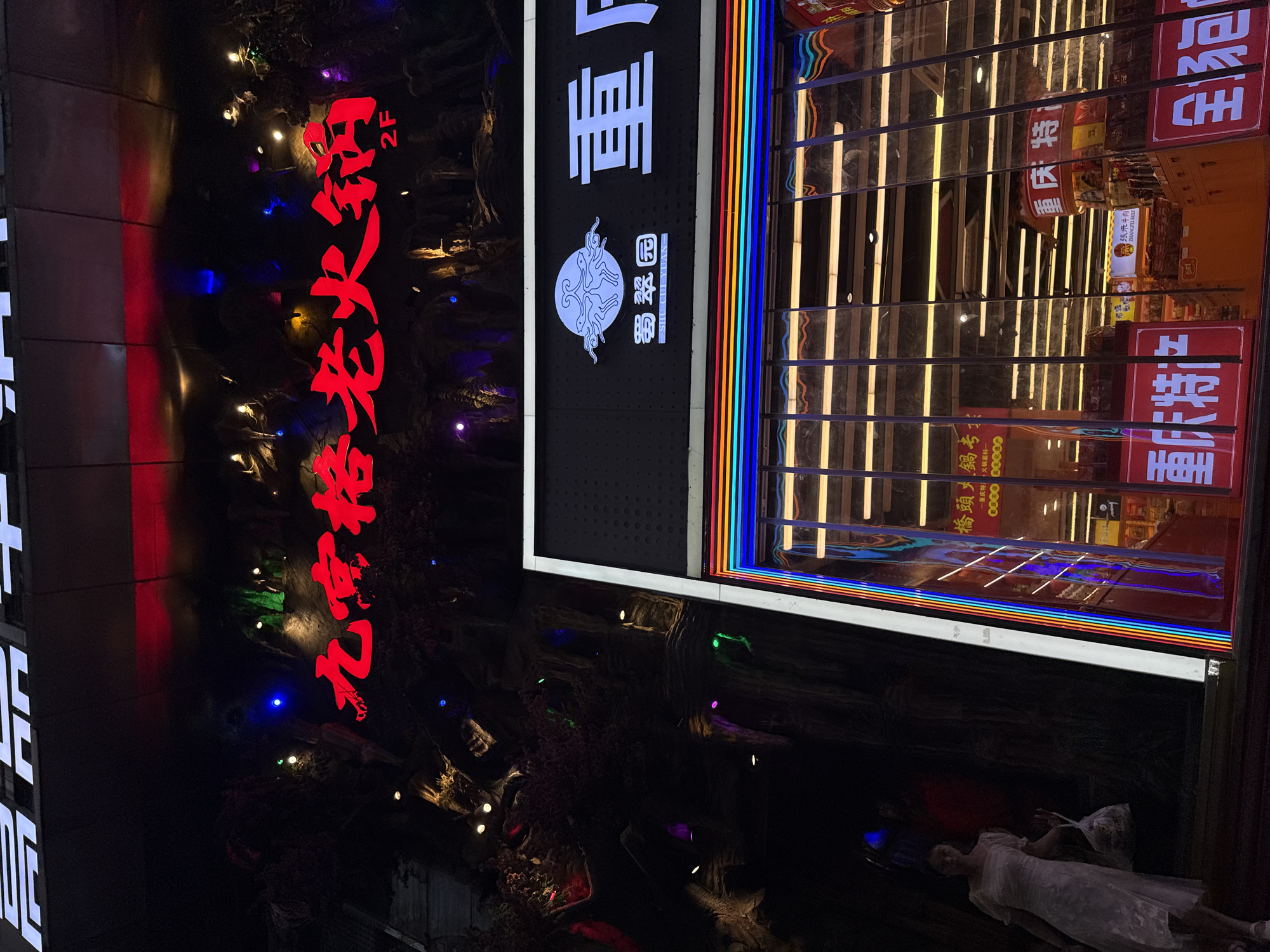 The image shows a building facade at night in the Yuzhong District of China. A large red neon sign with Chinese characters reads "九宫格老火锅" (Jiugongge Lao Huoguo), indicating a hot pot restaurant. Below this sign, "2F" is visible, denoting the second floor. To the right, another sign with Chinese characters, "重", is part of a larger business name. The building has a dark, reflective exterior, with what appears to be a faux rock or natural texture on the left side, illuminated by colorful lights. A woman in a white dress is walking towards the left of the frame, holding a shopping bag. The storefront below features large windows with colorful LED lighting along the frame. Inside the store, shelves stocked with products are visible through the reflection on the glass.