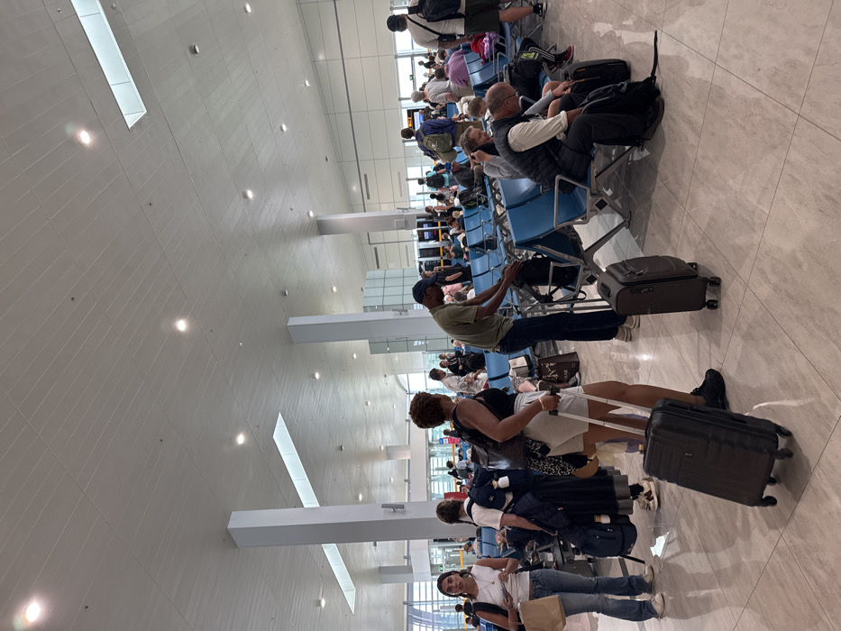 An indoor scene depicts a busy airport departure lounge at Abeid Amani Karume International Airport (ZNZ) in Zanzibar, Tanzania. The spacious terminal features a high white paneled ceiling with numerous recessed circular lights and large rectangular skylights. The floor is covered with polished light-colored tiles. Several white pillars support the ceiling.
Approximately 30-40 individuals, primarily adults, are present. In the immediate foreground to the left, a female with dark hair, wearing a white sleeveless top and blue jeans, stands holding a small brown paper bag. Beside her, a second female with dark hair, wearing a white top and dark long skirt, stands facing away from the camera, next to a large blue backpack. In the central foreground, a female with curly brown hair, dressed in a black tank top and light shorts, pulls a dark gray rolling suitcase. Behind her, a male wearing an olive green shirt, dark jeans, and a baseball cap bends over a black bag on a blue airport seat, a dark gray rolling suitcase at his feet. To his right, a male in a dark vest sits hunched on a blue seat, with a black backpack beside him, and a female is partially visible next to him.
Rows of blue, linked airport-style seating stretch across the midground, occupied by numerous passengers. More individuals are visible standing and sitting in the background, near what appear to be distant information screens or boarding gates. Most people are either seated and waiting or standing with their luggage. The scene is well-lit by both artificial and natural light from the skylights.