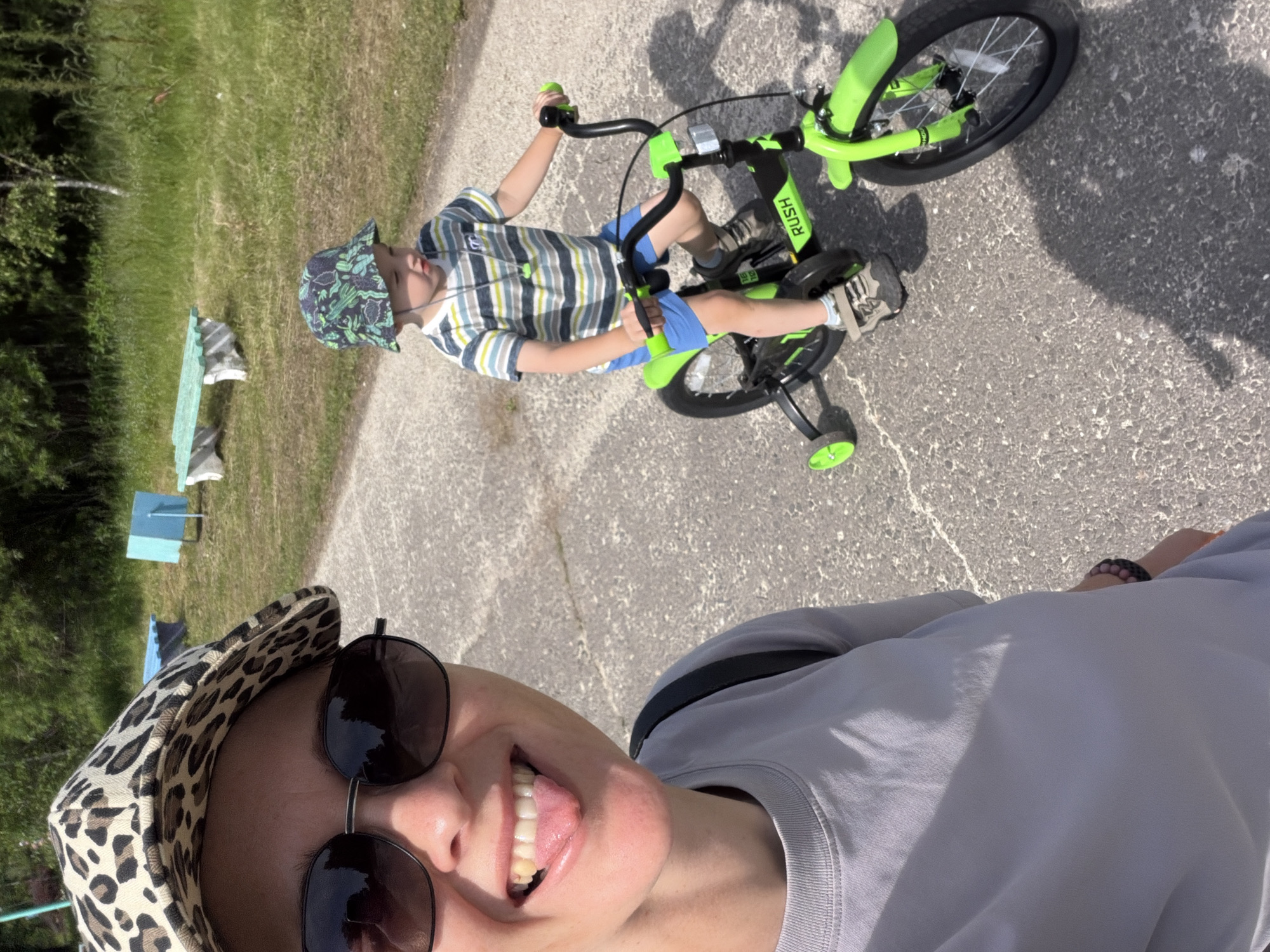 In Kul'dur, Russia, a woman with a leopard-print hat and sunglasses sticks her tongue out at the camera, her mouth wide open in a smile. She wears a gray t-shirt and a black backpack strap is visible over her shoulder. To her right, a young boy wearing a green bucket hat and a striped t-shirt rides a bright green bicycle with training wheels on an asphalt path. The boy is wearing blue shorts and sneakers. The background features a grassy area with trees and some outdoor furniture, including a light blue structure and a bench. The lighting suggests it is a sunny day.
