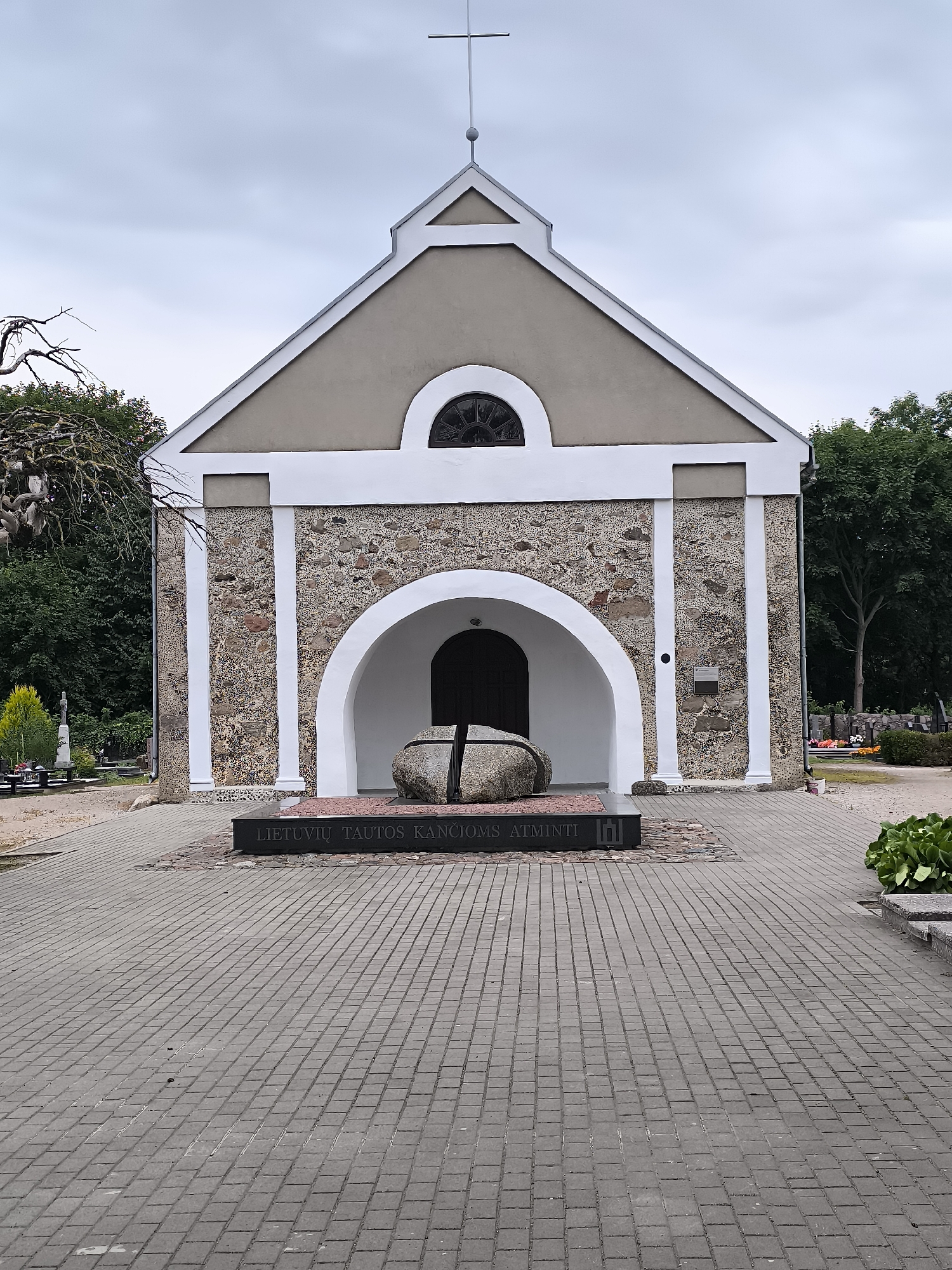 The image shows a view of the Kretinga cemetery in Kretinga, Lithuania. A small chapel with a stone facade and a grey roof stands prominently in the center. A cross adorns the peak of the chapel's roof. In front of the chapel, a large, split boulder rests on a dark stone plaque inscribed with Lithuanian text that translates to "Lithuanian Nation's Suffering and Hope." The ground in front of the chapel is paved with grey brickwork, and a path extends into the background, lined with gravestones and trees. The sky is overcast, suggesting a cloudy day. No people are visible in the image.