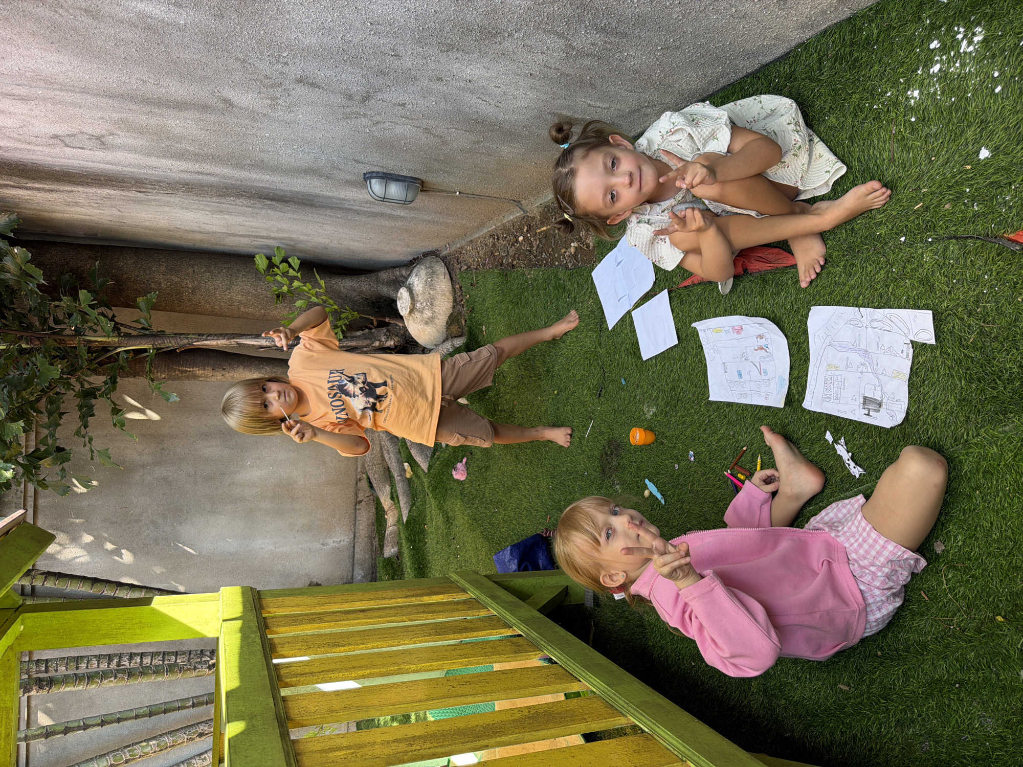In Kuta, Indonesia, three children are pictured in a backyard setting. A boy, standing, wears an orange t-shirt and brown shorts. A girl, sitting on the grass, is dressed in a pink jacket and pink and white checkered shorts. Another girl, also sitting on the grass, wears a light-colored floral dress. They are surrounded by drawings and art supplies scattered on the artificial turf. A wooden play structure is visible to the left, and a concrete wall runs along the right side of the image.
