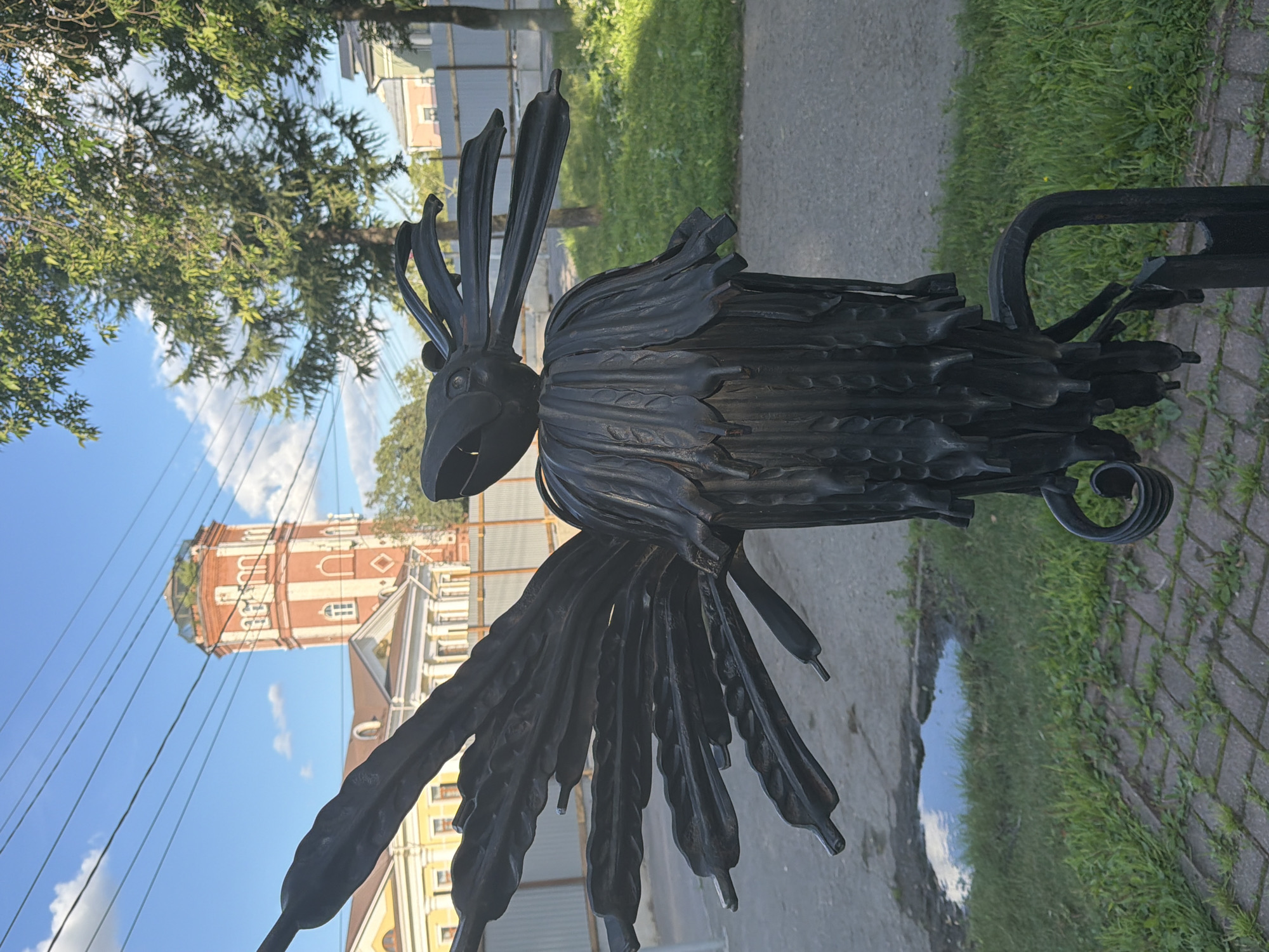 The image shows a dark metal sculpture of a stylized bird in the foreground, with its wings spread and beak open. The sculpture appears to be made of forged metal and is weathered. In the background, a red and white striped tower with a domed roof stands against a blue sky with scattered clouds. Several trees with green foliage are also visible, as well as a building with a yellow facade. The sculpture is located in a grassy area next to a paved pathway. The environment suggests an outdoor public space. The metal bird sculpture is situated in Vologda, Russia, outside the Vologda Regional Museum of Local Lore named after N.V. Vereshchagin, specifically near the observation deck of the former water tower.