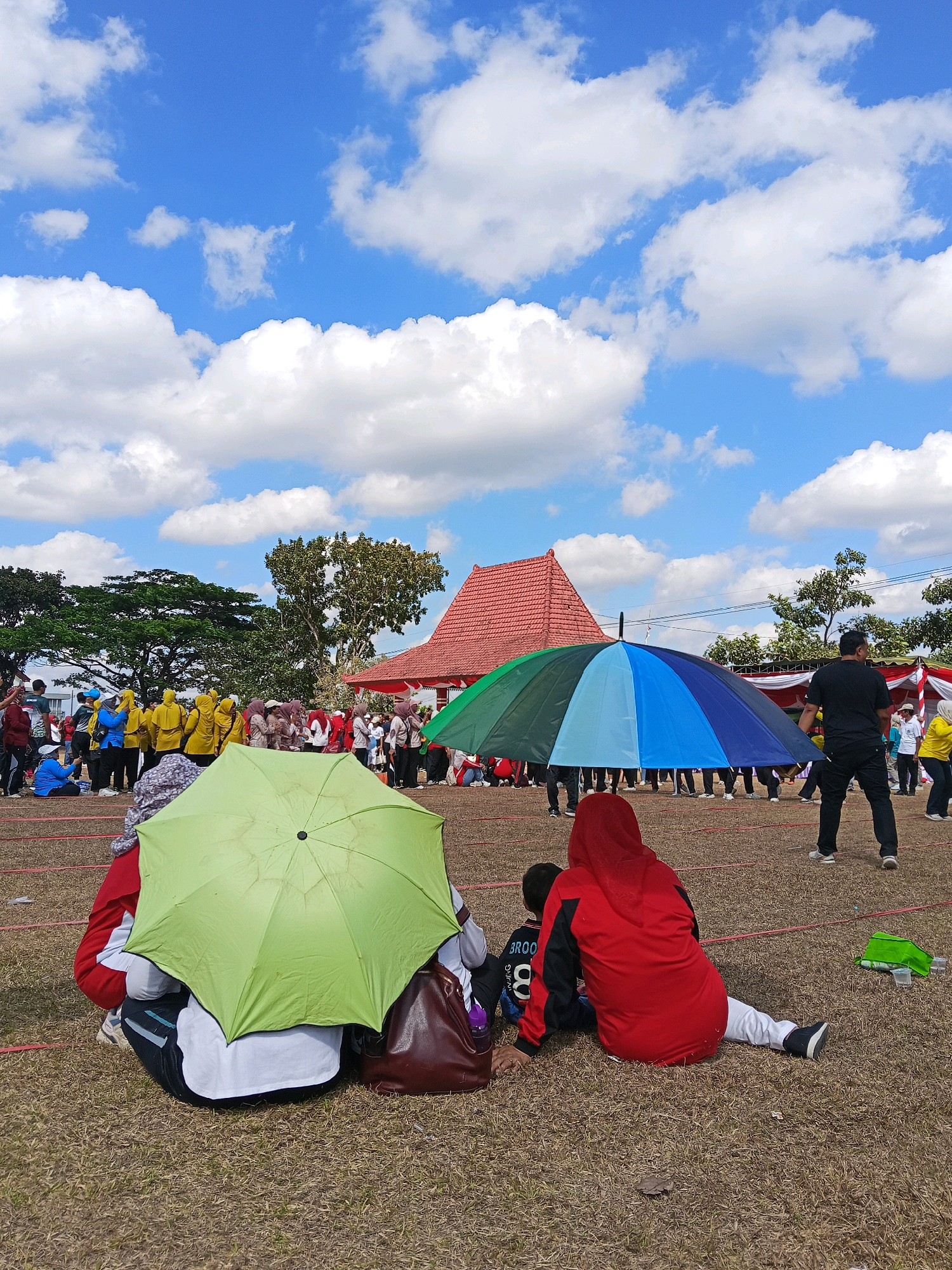 The image captures an outdoor event on a sunny day with scattered clouds in a bright blue sky. In the foreground, two people are seated on dry grass, one under a bright green umbrella and another wearing a red hijab and jacket. Several other individuals and groups are visible in the middle ground and background. A large, multi-colored umbrella, divided into green, blue, and dark sections, is being held up by several people.
In the background, there's a traditional Indonesian building with a red tiled roof, and trees line the perimeter of the open field. A group of people, some wearing yellow, are standing in an organized line. A man in a black shirt and pants is walking away from the camera towards the right side of the frame. The ground is covered with dry grass and demarcated by thin red lines, suggesting a sports field or event venue. The overall scene depicts a lively outdoor gathering or competition. The location is identified as Bendosewu, Indonesia.