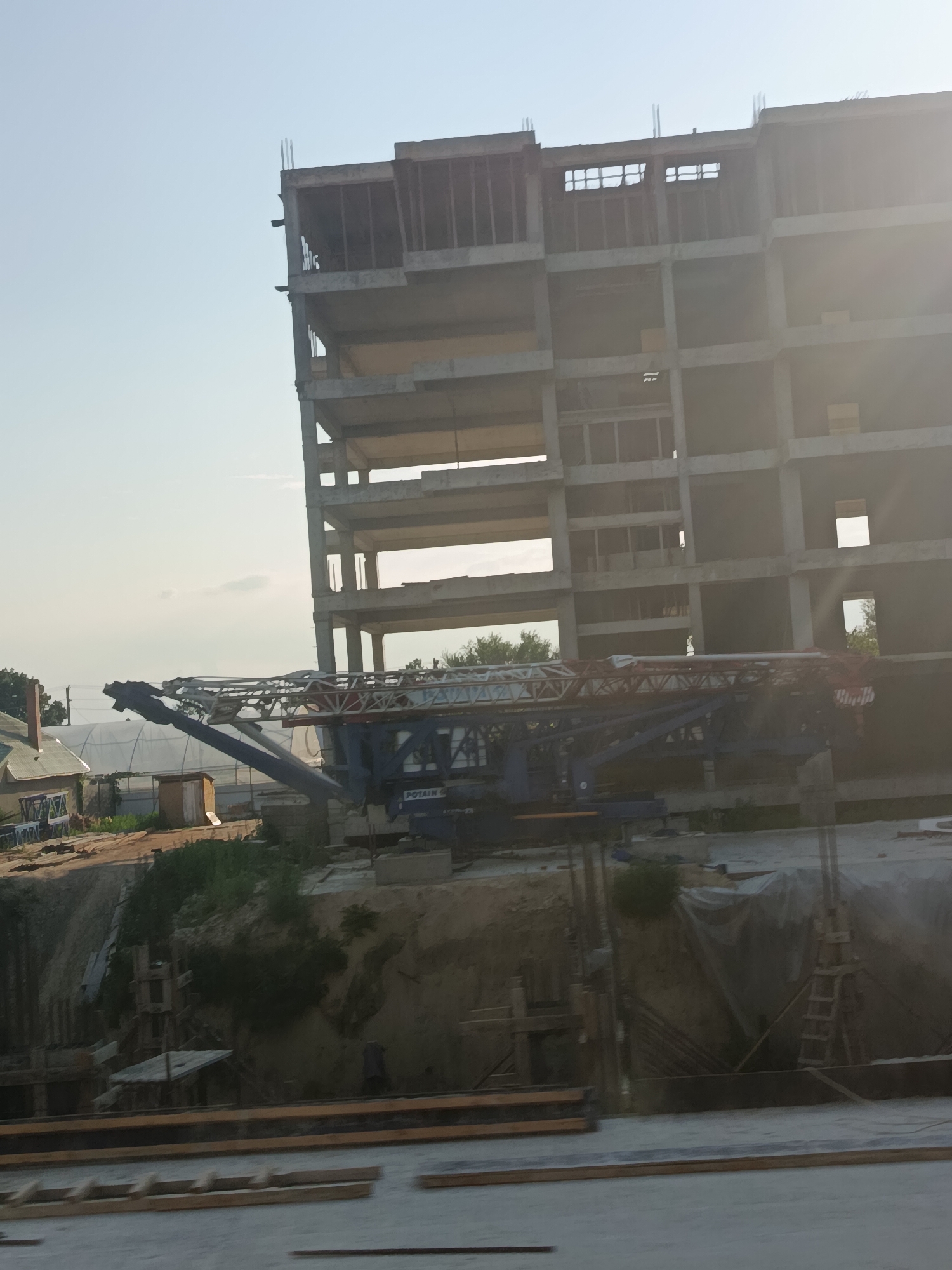 A multi-story concrete building under construction is visible, with rebar protruding from the top floors. A large construction crane, a Potain brand, is positioned in the foreground, its arm extended over the building. To the left of the building, a greenhouse-like structure and a small, wooden shed are present. The ground appears to be a construction site with exposed earth and some covered materials. The sky is clear and light blue. The location is identified as Cahul, Moldova.