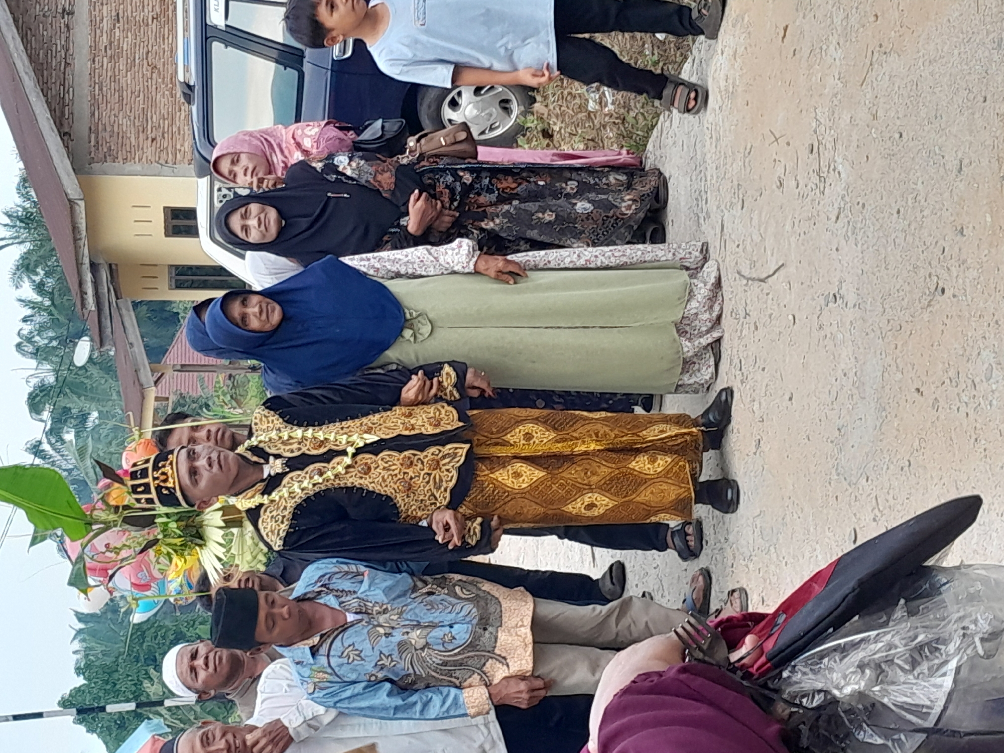 A group of people are gathered outdoors in what appears to be a village setting in Langga Payung, Indonesia. The central figures are a man and woman dressed in traditional attire. The man is wearing a black jacket with ornate gold trim, a gold necklace, and a patterned gold and brown sarong. He also wears a black, ornate headdress. The woman beside him is wearing a muted green long dress and a dark blue headscarf. To the left of the couple is a man in a light blue, patterned shirt and tan pants, and further left, another man in a white shirt and white cap. Behind them, several other women and children are visible, some wearing headscarves. In the background, a yellow building with a brown roof and a dark blue SUV are present. There are also some decorative elements, possibly part of a celebration, including balloons and leafy branches, on the left side of the image. The ground is paved with a rough, concrete-like surface. The lighting suggests it is daytime, with overcast skies.