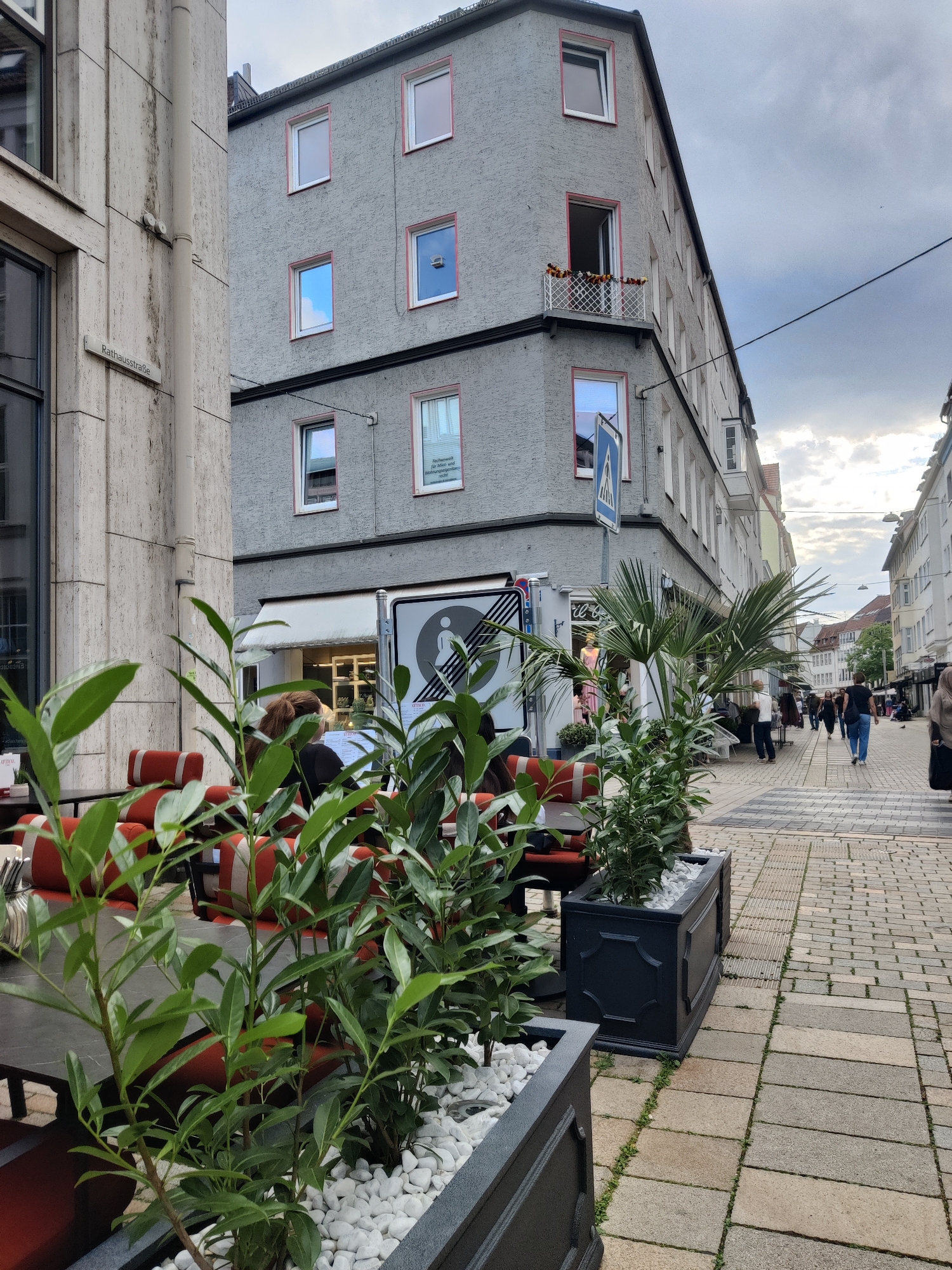 The image shows a street scene in Bielefeld, Germany. In the foreground, there are plants in dark grey planters with white stones. Behind the planters, there are tables and chairs with red and white cushions, suggesting an outdoor seating area for a cafe or restaurant.
The middle ground features a building with a grey facade and several windows. Some windows have pink trim. There is a street sign on the building that reads "Rathausstraße". A mobile concession stand or food truck with a white awning and a circular logo is parked on the sidewalk.
Further back, the street is paved with cobblestones and lined with buildings. Several people are walking along the sidewalk, some are shopping or dining. The sky is cloudy, indicating overcast weather.