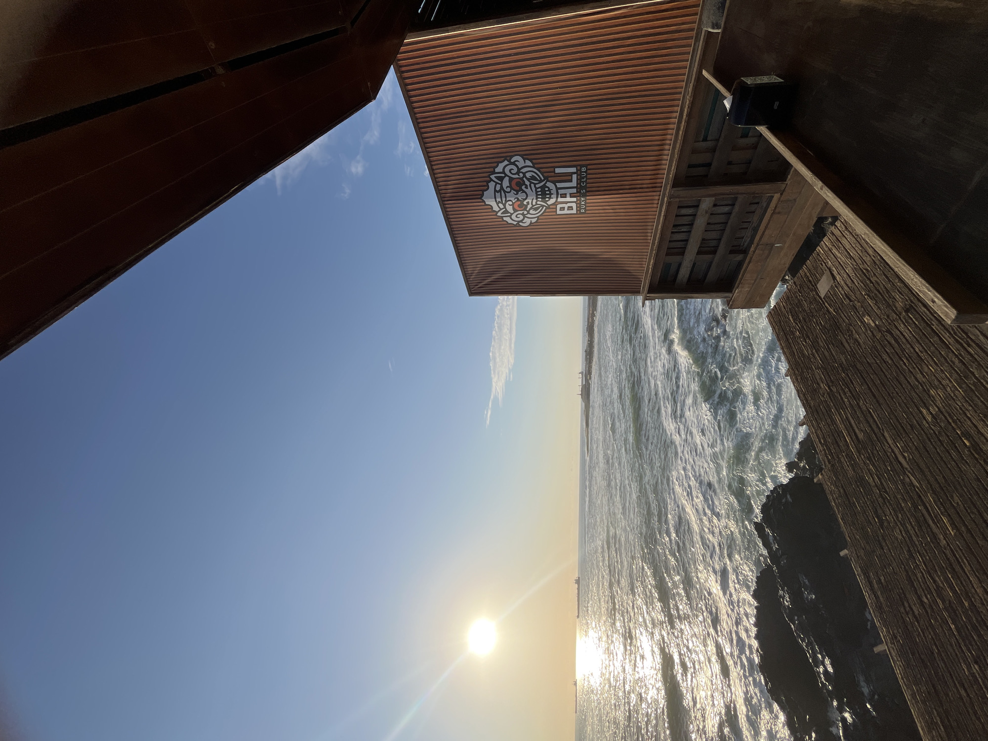 The image shows an ocean view in Arica, Chile. The sun is shining brightly over the ocean. A building with the logo "Bali Rugby's Club" is overhead. The ocean waves are crashing against the rocks. A partial view of two wooden buildings are visible. The sky is clear blue.