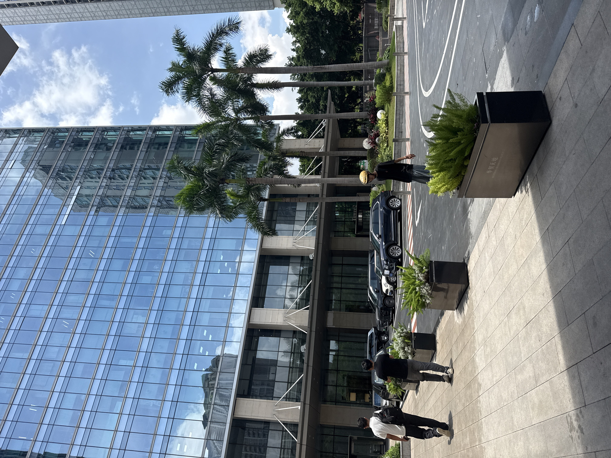 This image depicts the exterior of a modern, glass-fronted office building in Huizhou, China, likely a commercial or business district. The building features multiple stories of reflective blue glass windows, with the sky and surrounding elements mirrored on its facade. In front of the building, several parked cars are visible, including black sedans and an SUV, suggesting this is a parking area or drop-off zone.
Two individuals are walking on a paved walkway in the foreground, one wearing a white polo shirt and dark pants with a backpack, and the other in a black t-shirt and dark pants. Another person, wearing a yellow hard hat and a black t-shirt with grey pants, is walking in the mid-ground across a road marked with white lines. Palm trees are interspersed with the building's architecture, adding a tropical element to the urban landscape. Planter boxes with lush green foliage are placed along the walkway and near the road. The sky is partly cloudy with patches of blue. The overall impression is one of a well-maintained, contemporary urban environment.