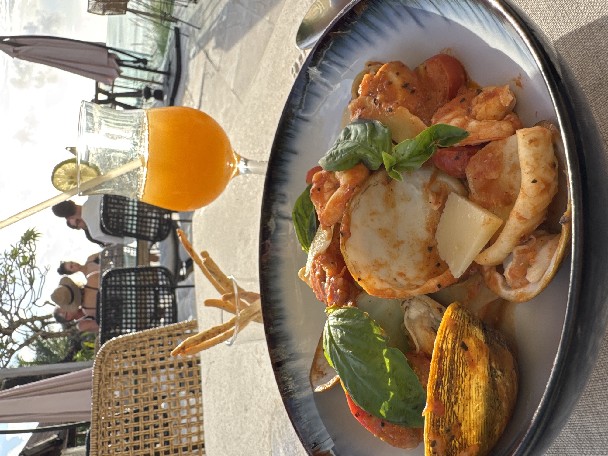 The image shows a table setting in Kuta Selatan, Indonesia. A plate of food dominates the foreground, containing seafood, tomatoes, potatoes, and basil. A glass of orange juice sits beside it, with a straw and lime slice. Breadsticks are also visible in a glass. In the background, blurred figures sit at tables with chairs, and a partially visible beach scene with umbrellas suggests a coastal location. The image is taken in daylight, with warm sunlight enhancing the colors.