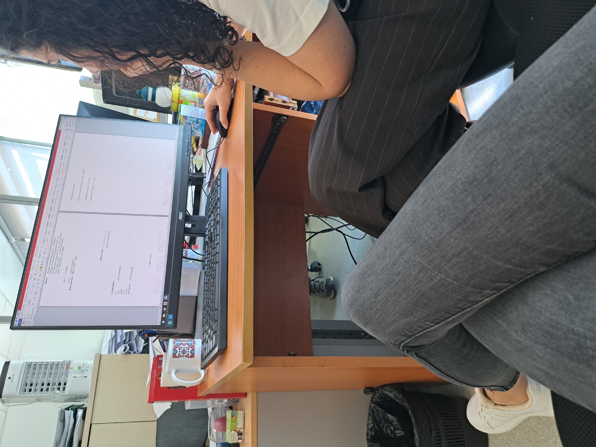 The image captures an individual engrossed in work at a computer within an office setting. The person, whose face is not visible, has dark, curly hair and is seen from behind and the side, wearing a white t-shirt and dark, pinstriped trousers. They are seated at a wooden desk, operating a computer mouse with their right hand while focused on an AOC brand monitor. Their legs, clad in grey trousers/jeans and white Adidas sneakers, are visible in the foreground and under the desk.
The computer screen displays a document, predominantly in Bulgarian, which appears to be a project proposal or technical report. Key visible text includes "Обект" (Object) detailing a project for "Столична Община" (Sofia Capital Municipality), "Част: Геодезия" (Part: Geodesy), and mentions "Възложител: ЕРМ Запад АД" (Client: ERM Zapad AD) and "Изпълнител: "МАПЕКС" АД" (Contractor: "MAPEX" AD). The document is dated "София 2026 г.", suggesting a future-oriented project or draft.
The desk is equipped with a black keyboard and a white mug featuring a colorful, intricate pattern, possibly traditional Bulgarian. To the left, a small fan or cooler unit is visible. The immediate surroundings suggest a typical office environment, with various papers and office supplies. The overall lighting indicates daytime. The visible text confirms the location context of Sofia, Bulgaria.
