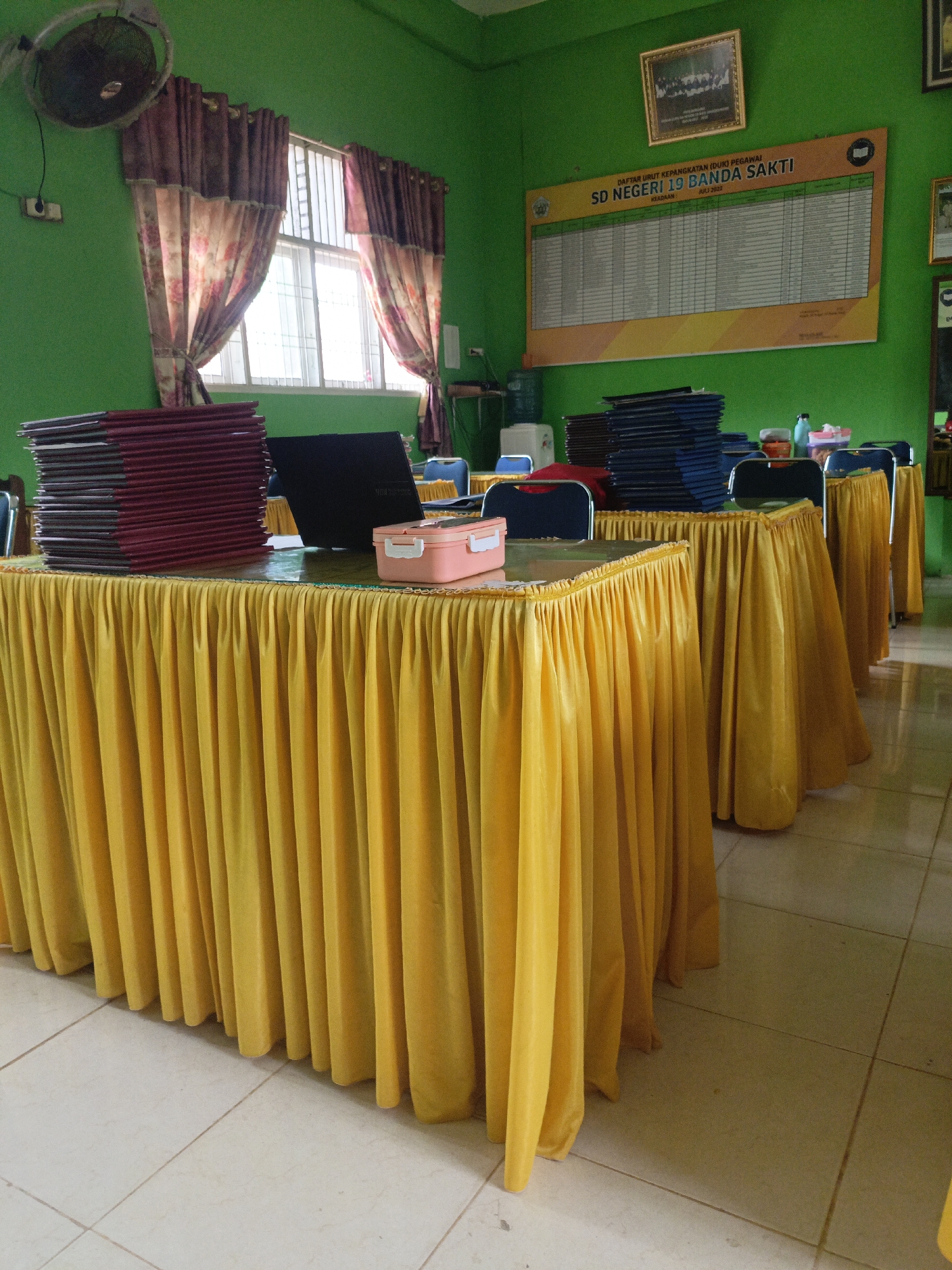 The image depicts the interior of a classroom or meeting room in a school in Lhokseumawe, Indonesia. The walls are painted a bright green, and a large bulletin board displaying a staff list for "SD NEGERI 19 BANDA SAKTI" is mounted on one wall. A framed group photograph hangs above the bulletin board.
In the foreground, a long table draped with a yellow tablecloth is visible. On the table, stacks of maroon and dark blue folders or binders are neatly arranged. A black laptop and a pink and white rectangular container are also present on the table.
To the left of the table, a wall-mounted fan with metal blades is attached to the green wall. A window with white blinds and patterned curtains, featuring floral motifs in pink and brown, allows natural light into the room.
In the background, other tables draped with similar yellow tablecloths and chairs with blue seats and black frames are arranged in a row. Bottles of liquid and other small containers are placed on these tables. The floor is tiled with light-colored square tiles. The overall impression is one of an organized and functional space within a school environment.