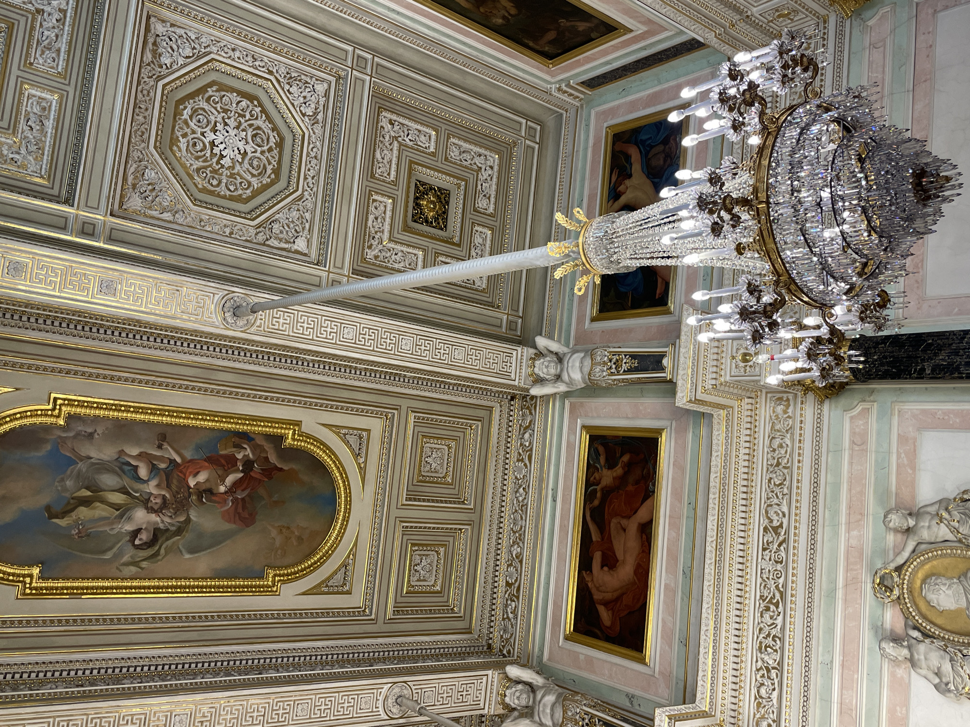 The image shows a ceiling with a large chandelier, likely inside a building. The ceiling is divided into rectangular sections, with some containing paintings and others featuring ornate plasterwork in gold and white. The paintings depict human figures, possibly angels or mythological characters. The chandelier is made of crystal and metal, and appears to be illuminated. The location is unknown, but the style of architecture and decoration suggests a historic or palatial setting in an unknown city.