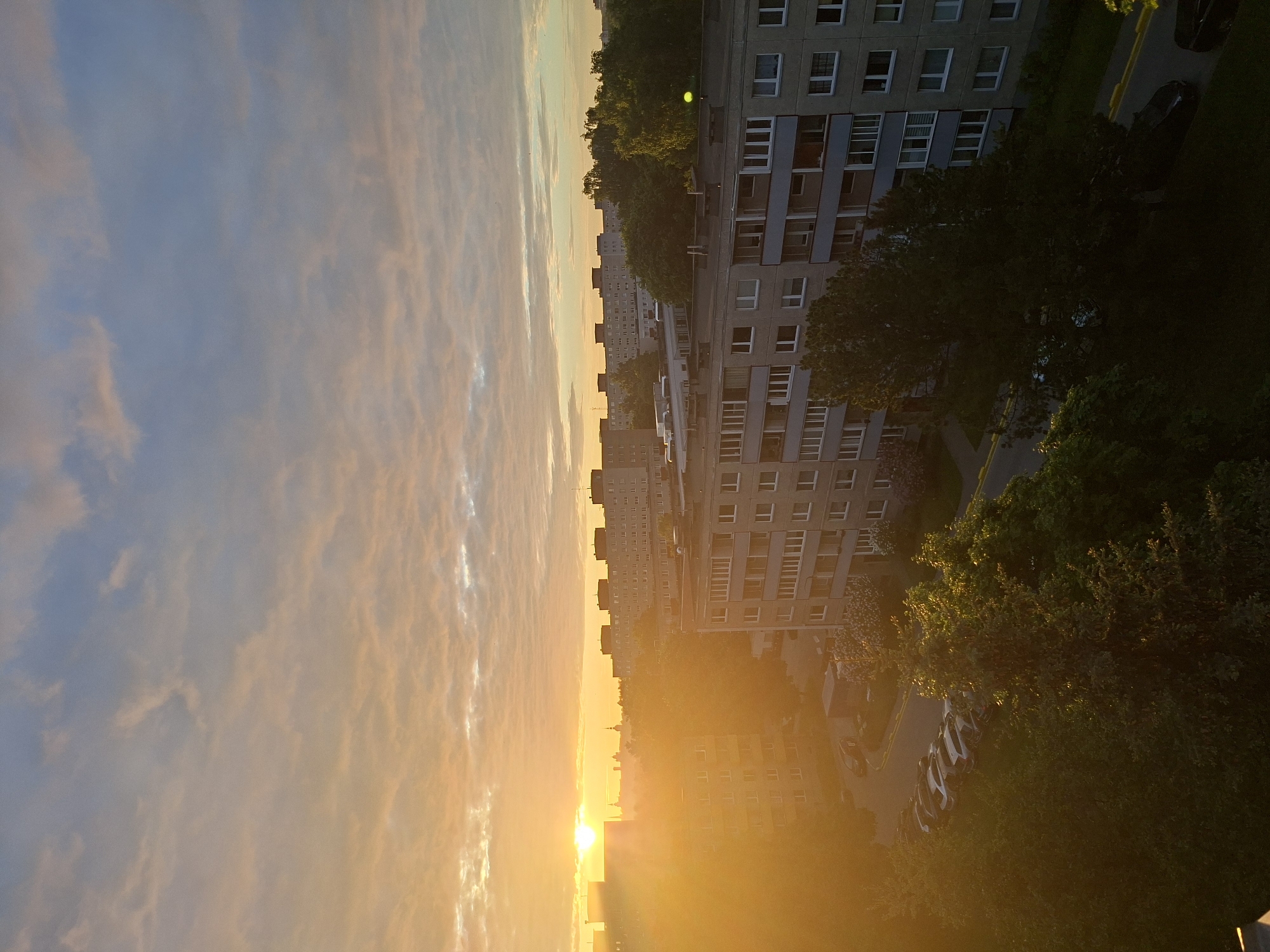 The image depicts an urban or suburban landscape at either sunrise or sunset, captured from an elevated vantage point. The sky dominates the upper and left portions of the frame, showcasing a gradient from soft blue-grey to an intense, warm orange and yellow near the horizon. Streaky, illuminated clouds are visible across the sky.
The sun is a brilliant, concentrated orb of light located just above the horizon on the right side, casting a strong, golden glow across the scene and creating a distinct lens flare.
Below the sky, a series of multi-story residential or apartment buildings stretch across the middle ground. The most prominent building on the right is a grey structure with numerous rectangular windows, some appearing to have blinds or open panes. Further back, similar buildings are silhouetted against the bright horizon.
Dense green trees and foliage are abundant, particularly in the mid-ground and foreground on the right. Some trees are brightly illuminated by the low sun, while others are cast in deep shadow, adding depth and contrast. Hints of roads or parking areas, with what appear to be parked cars, can be faintly discerned among the trees and buildings in the lower right section of the image.
The scene conveys a peaceful atmosphere, with no people or specific activities visibly taking place. The overall impression is one of natural beauty and urban architecture bathed in the dramatic light of dawn or dusk. There is no visible text in the image.