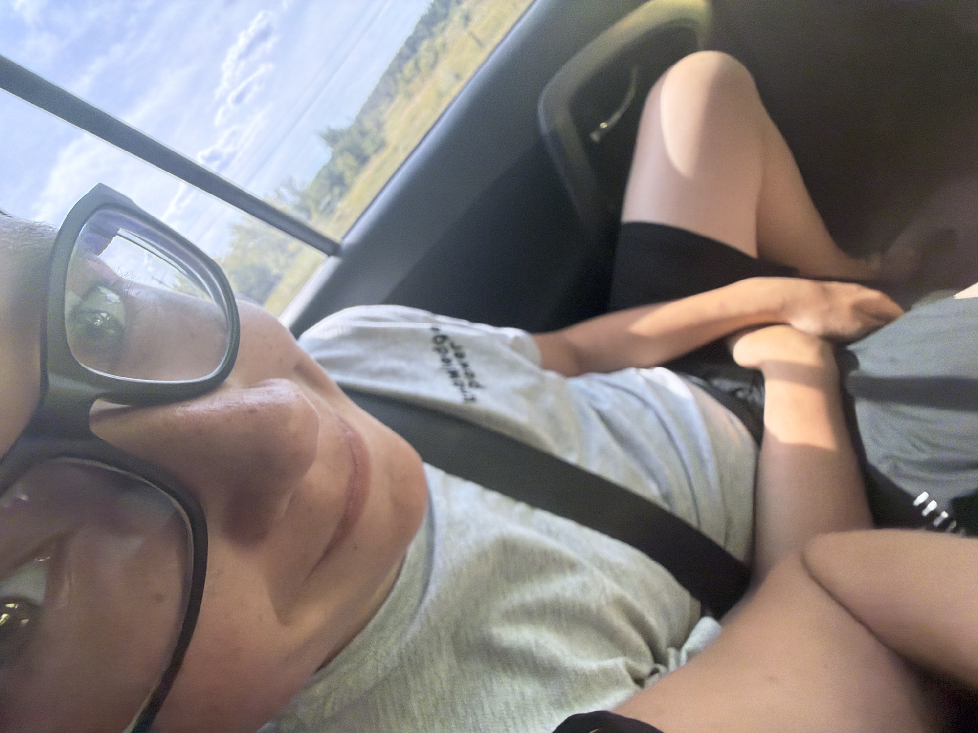 In Kruglovka, Russia, a close-up shot captures a person wearing glasses and a light-colored t-shirt with a black seatbelt crossing their chest. The t-shirt has text printed on it that is partially visible. The person is seated inside a vehicle, with the door and window visible in the background, showing a glimpse of the outdoor environment with trees and a cloudy sky. The lighting suggests it is daytime.