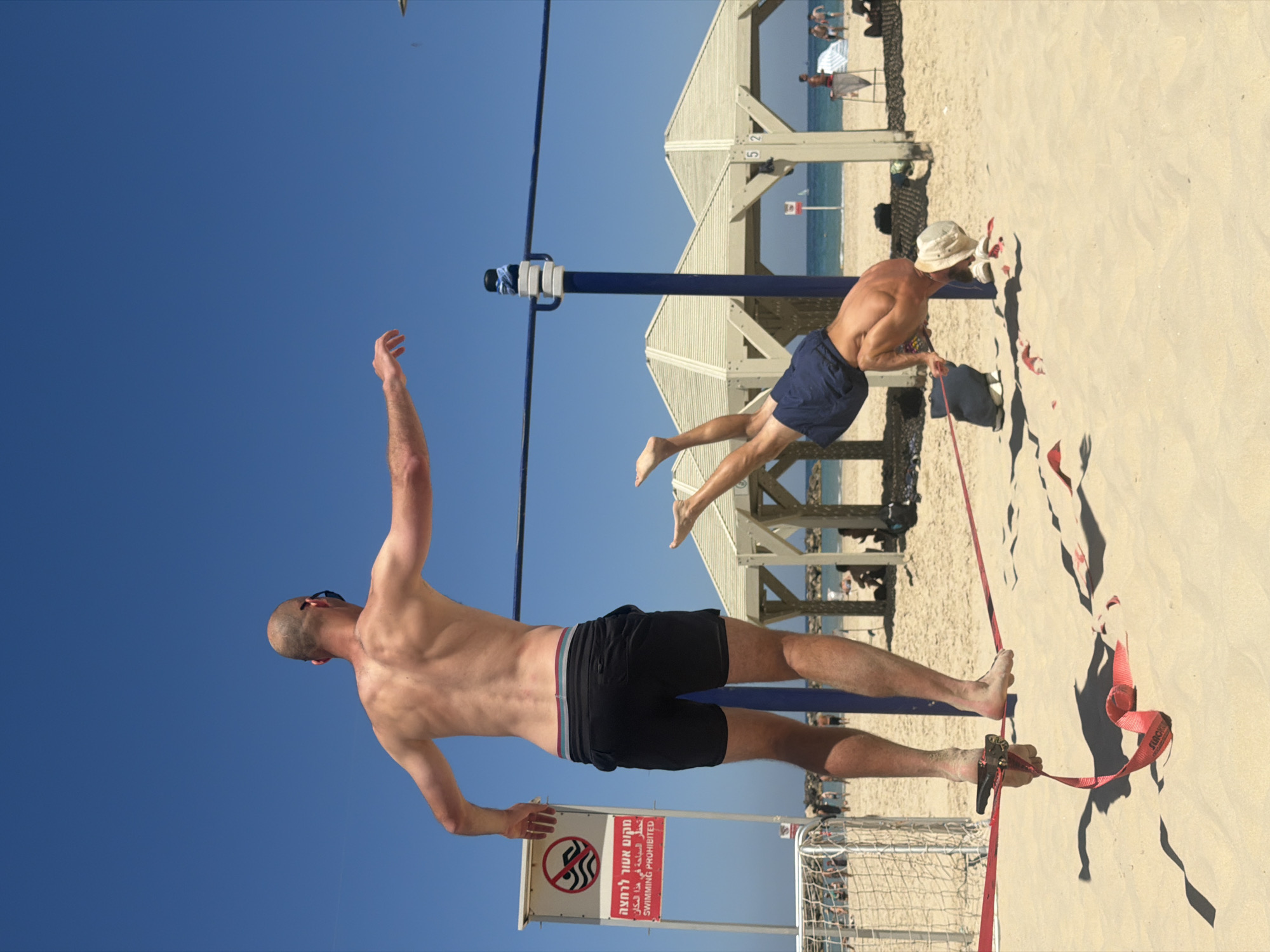 This dynamic shot captures a sunny day on a beach in Tel-Aviv, Israel, likely depicting a beach volleyball game or similar recreational activity. Two men are prominently featured on the sand.
In the foreground, a shirtless man with a shaved head and sunglasses is positioned on the left side of the frame, wearing black swim shorts. His right arm is extended upwards and backward, suggesting he is about to hit or serve a ball, or perhaps reaching for something overhead. He stands barefoot in the sand.
To the right, a second shirtless man in blue swim shorts and a light-colored hat is seen near a beach volleyball net. He is bending over, possibly adjusting equipment or retrieving something from the sand. His body is angled towards the right, with bare feet visible.
The scene is bathed in bright daylight under a clear, vibrant blue sky. The sandy beach stretches into the background, where the Mediterranean Sea is visible under a distant horizon. Several beach shelters or pergolas, characteristic of Israeli beaches, are scattered in the midground, with faint figures of other beachgoers near them. A blue net pole, part of a beach volleyball setup, stands between the two men.
Notable objects include a red strap or slackline lying on the sand in the bottom right, with "SLACK" visible on it, possibly for another activity or marking. Below the man in black shorts, a white sign with a red border and a "no swimming" icon is posted. The sign displays text in Hebrew, Arabic, and English, explicitly stating: "מקום אסור לרחצה" (Swimming prohibited place), "تحظر السباحة في هذا المكان" (Swimming is prohibited in this place), and "SWIMMING PROHIBITED." Numbers "5" and "2" are also visible on a pole structure.