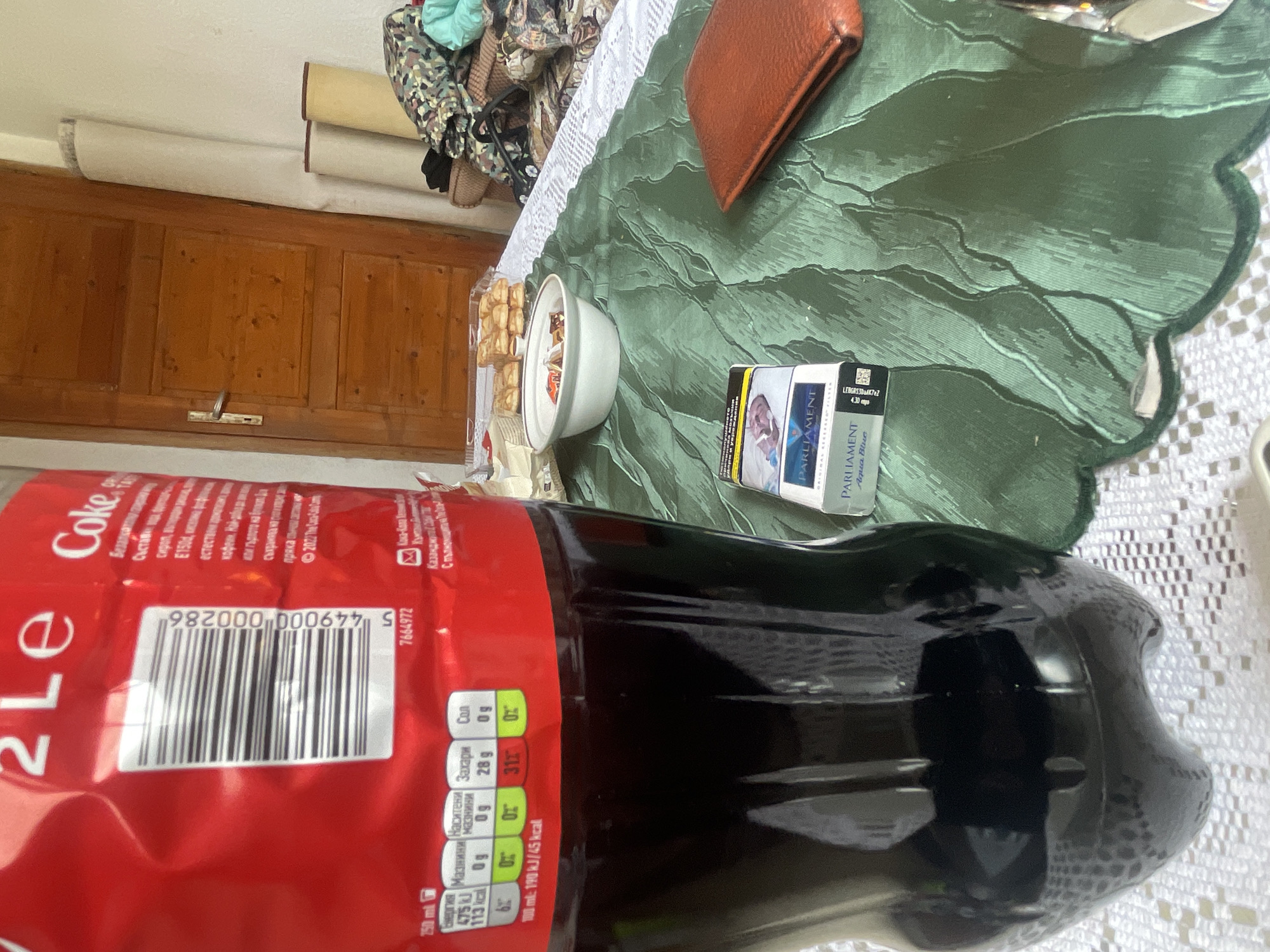The image shows a table setting in what appears to be a home in Kozarevo, Bulgaria. On a green leaf-patterned tablecloth, there's a pack of "Parliament Aqua Blue" cigarettes, a bowl containing small, possibly sweet, pastries, and a brown leather wallet. Dominating the foreground is a large 2-liter bottle of Coca-Cola. The table appears to be situated near a wooden door, suggesting a domestic interior. There are no people visible.