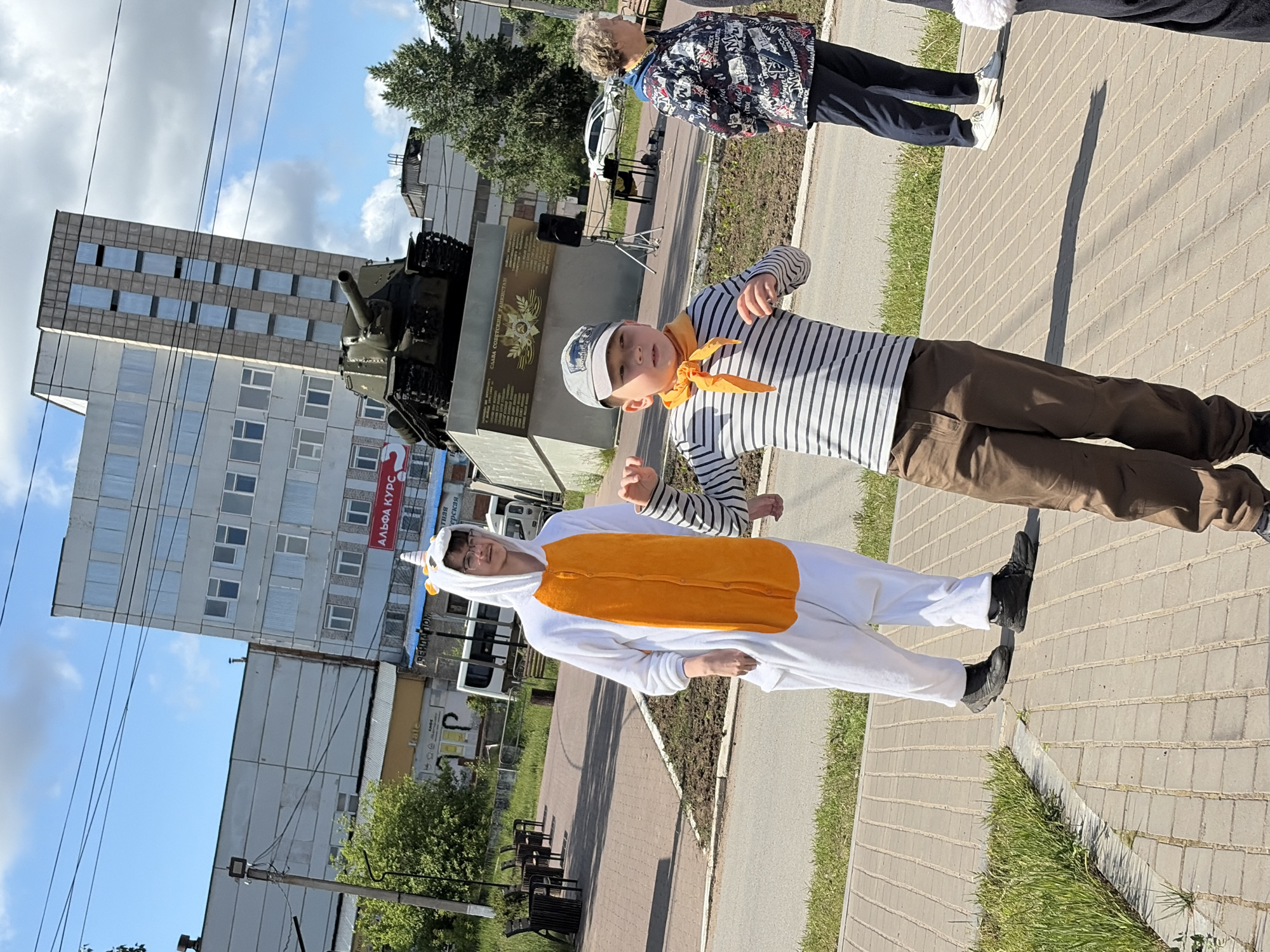 Two boys are standing on a paved sidewalk, in front of a war memorial. One boy is wearing a white unicorn onesie with an orange torso. The other boy is wearing a gray and white striped shirt, brown pants, and a gray cap, with an orange scarf tied around his neck. Behind them, a war memorial featuring a tank and a stone wall with inscriptions is visible. In the background, a tall, rectangular building and trees are partially visible against a blue sky with clouds. Another person is walking in the background. The scene is located in the city of Chaikovsky, Russia.