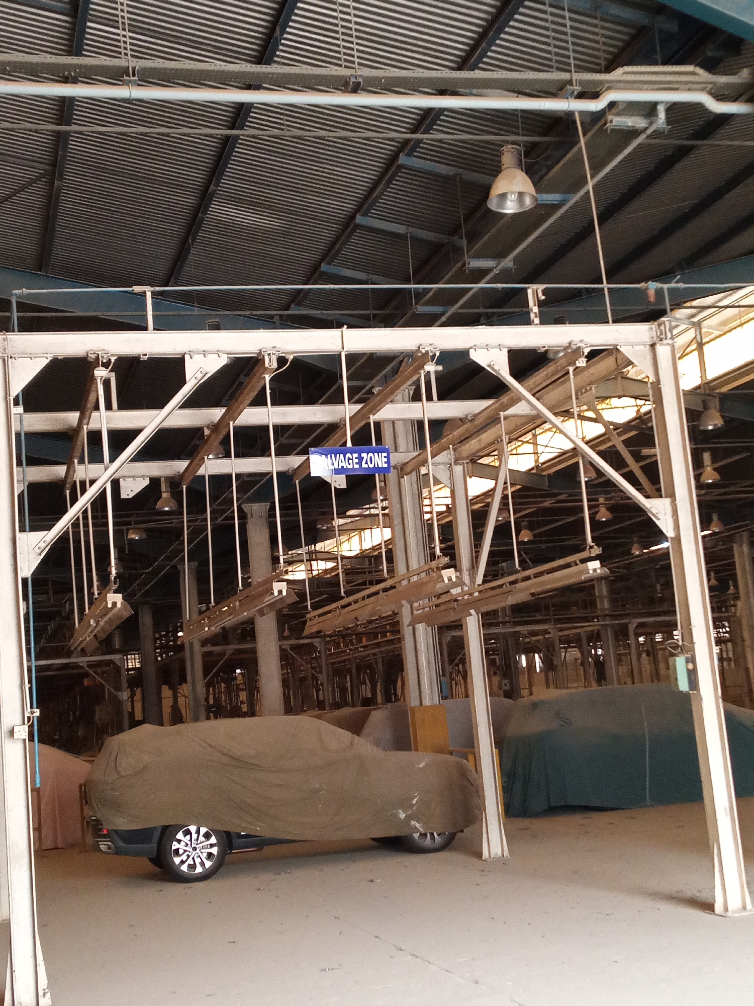 The image shows an indoor scene, potentially a large warehouse or factory in Kakuri Gwari, Nigeria. Several cars covered in protective sheets are parked inside. Above the cars is a complex network of metal support beams, pipes, and hanging lights. A blue sign with the words "SALVAGE ZONE" is visible among the overhead structures. The floor is concrete, and the overall lighting is somewhat dim. There are no people visible in the frame. The environment appears to be industrial and perhaps used for storage or vehicle processing.