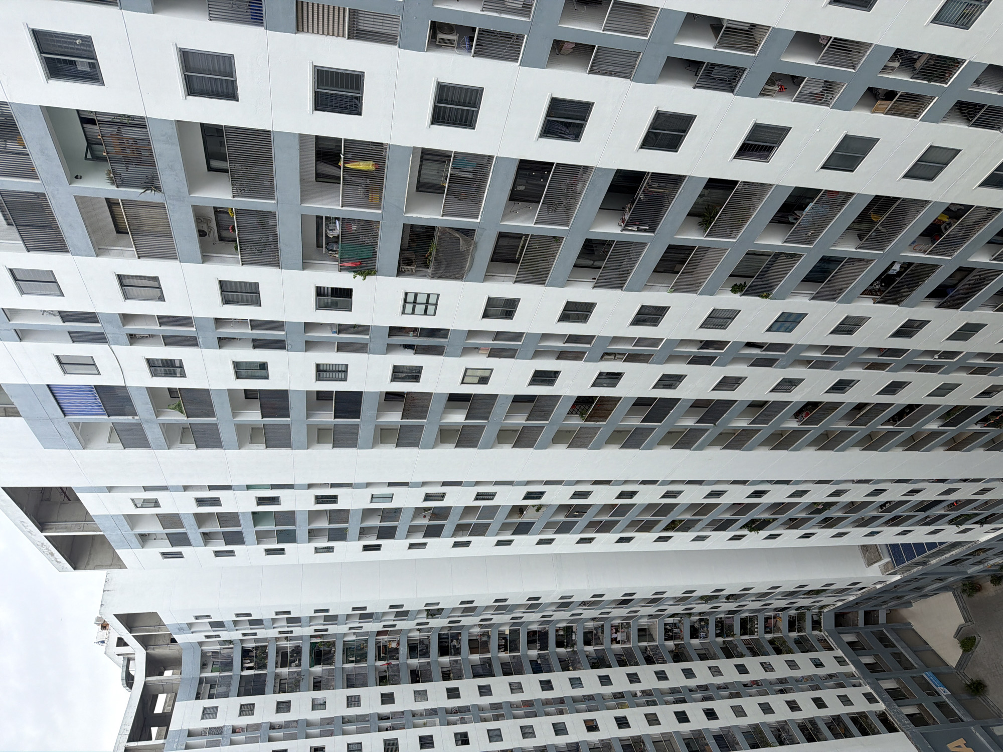 The image shows a high-angle view of a residential apartment complex in Nha Trang, Vietnam. The buildings are tall and feature a modern design with white and grey facades, balconies with metal railings, and numerous windows. The architecture suggests a dense urban environment with multiple interconnected structures, some of which appear to be under construction or undergoing renovation due to visible scaffolding and exposed concrete. The balconies display everyday life with laundry hanging out to dry, potted plants, and various personal items, indicating the presence of residents. The overall impression is one of a large-scale, multi-story housing development in a coastal city, given the overcast sky which is typical of Nha Trang's climate. There are no visible people in the image, but the signs of occupancy are evident.