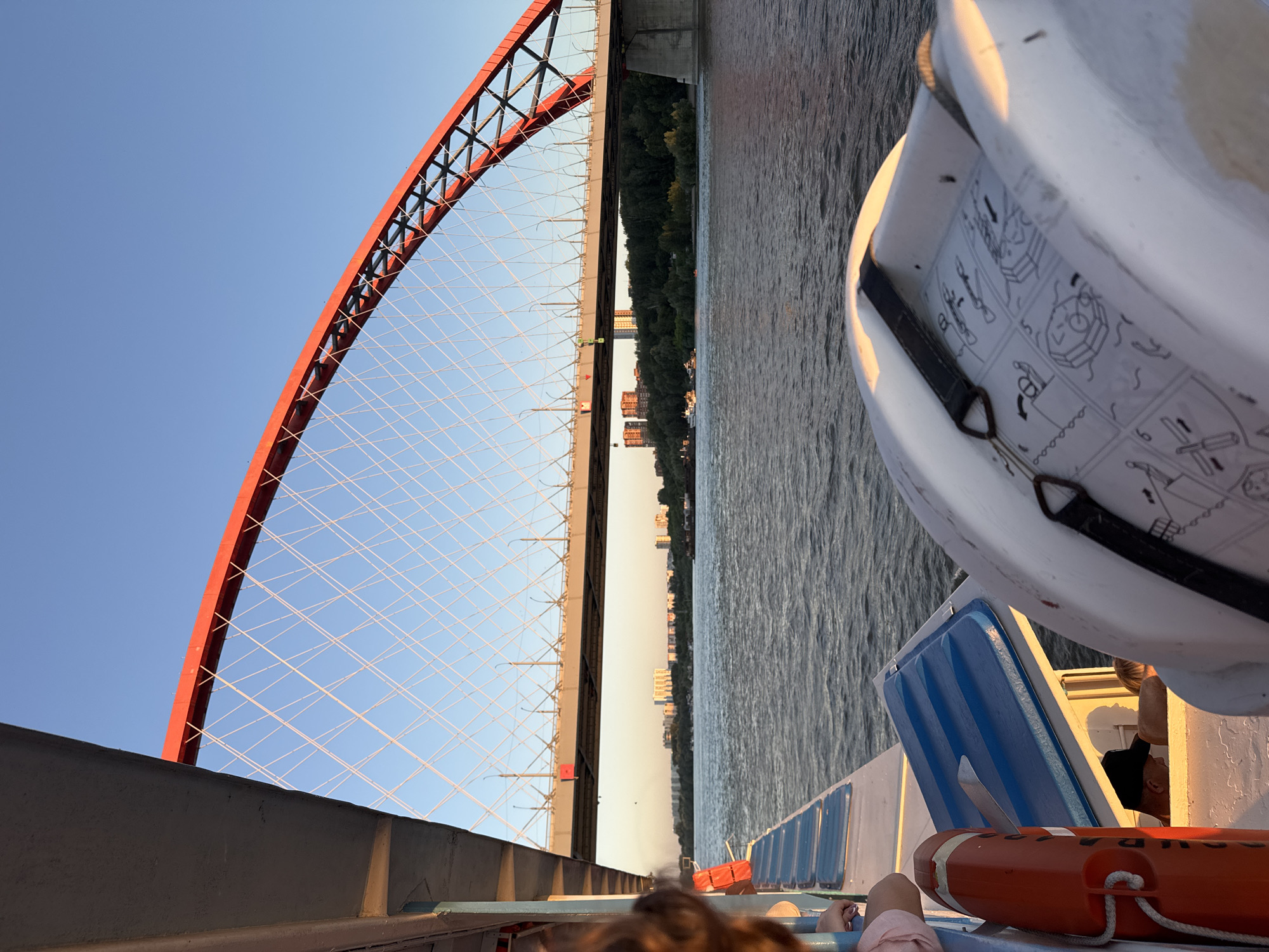 The image captures a view from a boat on the Ob River in Novosibirsk, Russia, with the Bugrinsky Bridge dominating the background. The bridge's distinctive red arch and suspension cables are illuminated by the setting sun, casting a warm glow on the scene. In the foreground, the side of the boat is visible, featuring blue seating and a life preserver. A white life raft with illustrated instructions for use is prominently displayed. The river's surface is rippled, reflecting the clear blue sky. Beyond the bridge, the Novosibirsk skyline can be seen with buildings nestled amongst a green, tree-lined shore.