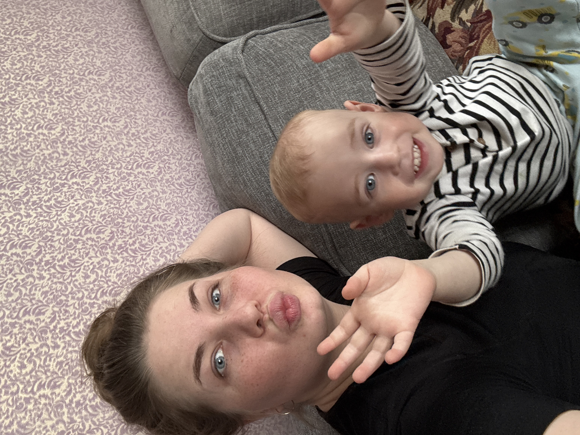 A selfie-style image shows a woman and a young child lying down. The woman is on the left side of the image, wearing a black shirt and pouting. Her hair is pulled back, and she has fair skin with freckles. Her right hand is raised, palm facing forward. The child is on the right side of the image, wearing a striped black and white long-sleeved shirt. The child is smiling and has their right hand raised in a similar manner to the woman. They are lying on a grey upholstered couch and what appears to be a patterned bedspread. The location might be Уральское, Russia, but that cannot be verified from the image itself.