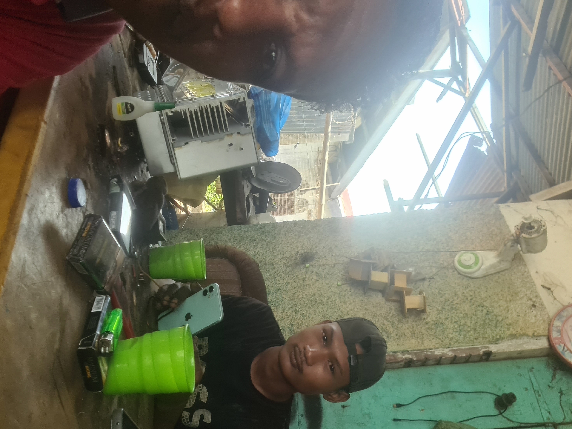 Two individuals are present in a workshop setting, likely in Lhokseumawe, Indonesia, suggested by the style of construction and the ambient conditions. The man on the left is partially visible, his face close to the camera. The man on the right wears a black t-shirt and a dark baseball cap. He is holding a light-colored smartphone and looking towards the camera.
On a cluttered wooden table between them are several objects: two bright green plastic cups, a pack of cigarettes labeled "MAGNUM BINTANG," a green lighter, and a white bottle with a green cap, possibly glue. There's also a piece of electronic equipment with fins, appearing to be a heatsink, and other assorted items suggesting repair work.
The environment is rustic and somewhat exposed. The roof structure above appears to be corrugated metal with visible wooden beams, some of which seem damaged or incomplete, suggesting a basic or under-construction shelter. The wall behind the man on the right has a textured finish and a light fixture is visible on it. A green wire hangs from the ceiling. The lighting is bright, indicating daylight.