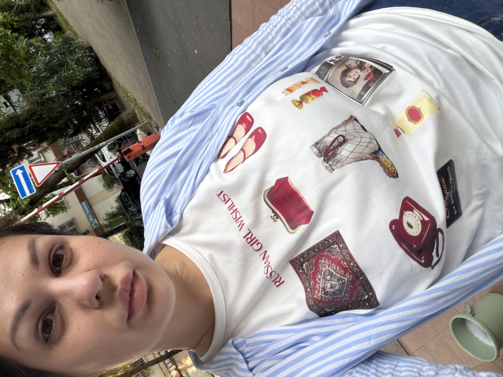 In Sochi, Russia, a young woman with dark hair and light skin is captured in a selfie taken outdoors. She is wearing a white t-shirt with various graphic prints, including the text "RUSSIAN GIRL WISHLIST," a red purse, a patterned rug, red high-heeled shoes, a vintage-style telephone, a pair of fishnet stockings, a bottle of perfume, a candy, and a picture on a television screen. Over the t-shirt, she wears a light blue and white striped button-down shirt, partially open. The background shows a street scene with a sidewalk, trees, buildings, and traffic signs, suggesting an urban environment. A red and white barrier is visible, along with parked cars and a motorcycle.