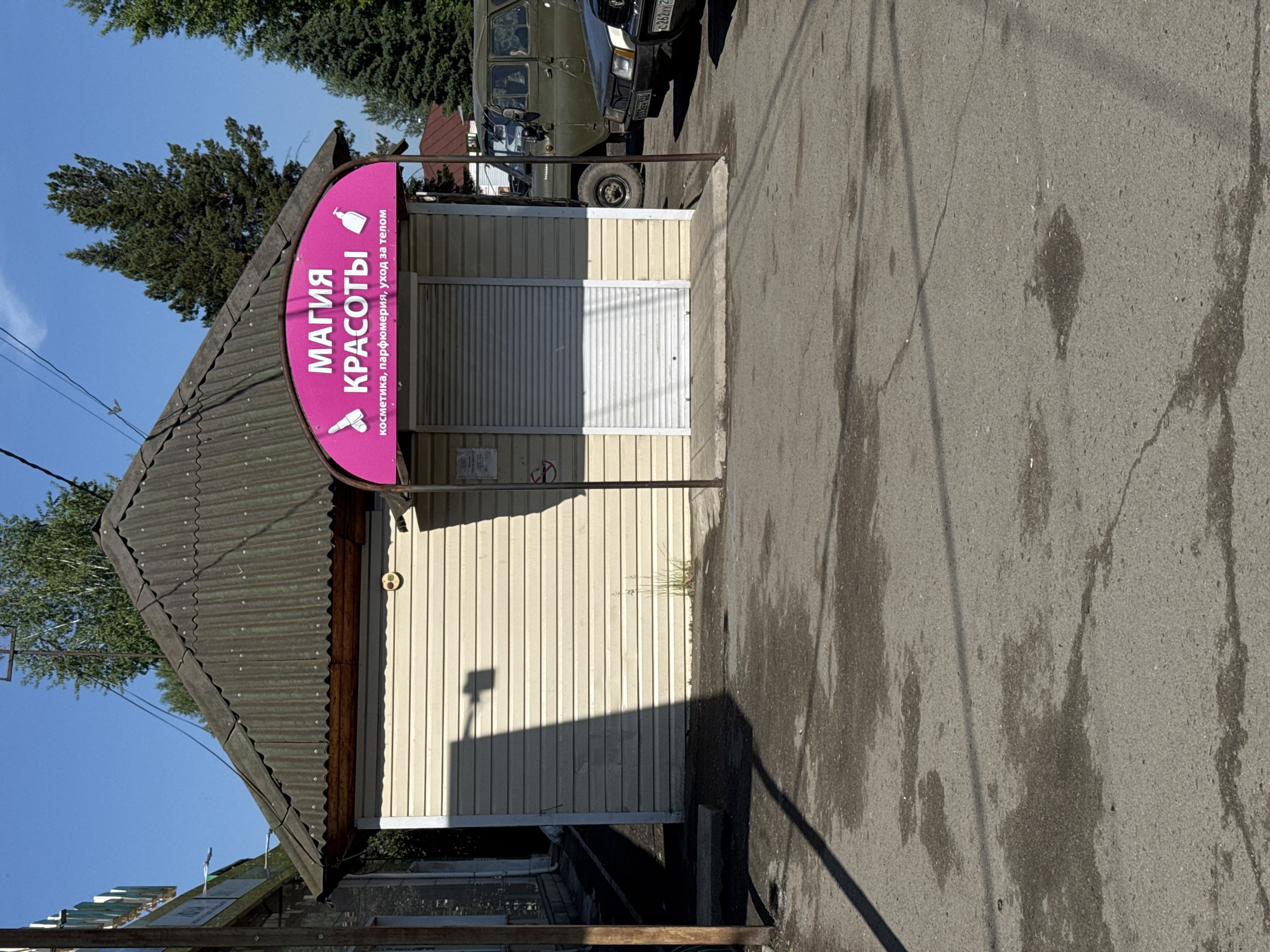A small, single-story commercial building with a corrugated metal roof is situated on an asphalt surface in Soloneshnoye, Russia. The building's exterior is clad in horizontal, light-colored siding, and it features a closed roll-up door. A pink, arched sign above the door displays Russian text that translates to "Magic of Beauty" and lists services like "cosmetics, perfumery, body care." Two small icons, resembling a lipstick and a soap dispenser, are also present on the sign.
Adjacent to the building, a dark green utility vehicle and a dark car are parked. The environment appears to be outdoors on a sunny day, with a clear blue sky visible, and shadows cast by the building and a nearby pole. The ground shows signs of dampness and cracking asphalt. In the background, various trees, including pines, are scattered.