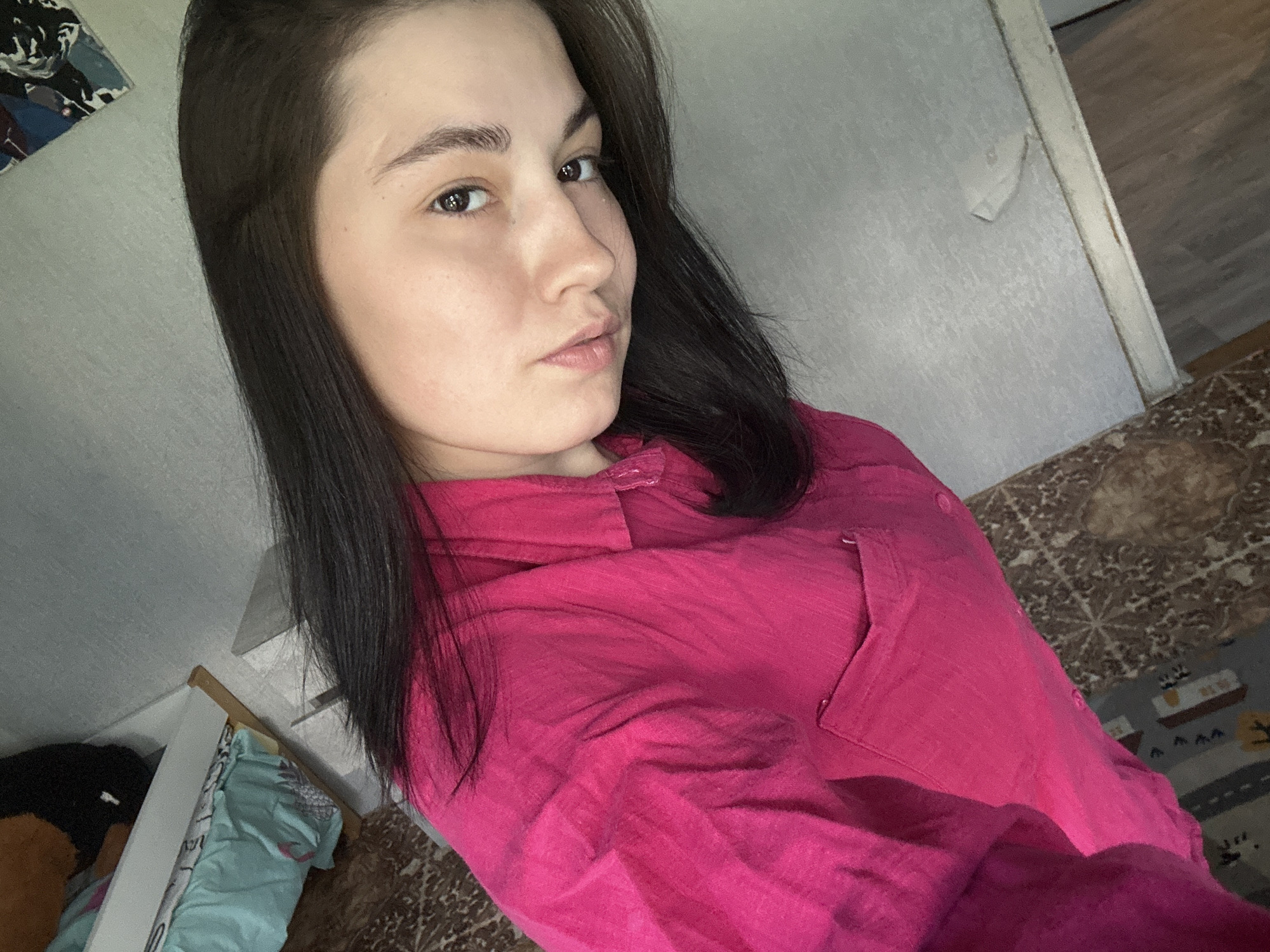 The media file shows a young woman taking a selfie in what appears to be a domestic setting. The woman has dark hair and is wearing a bright pink button-up shirt. She is looking towards the camera with a neutral expression, her gaze directed slightly off-center. The background is somewhat cluttered, with a wall that shows some peeling wallpaper and a doorway leading to another room with wooden flooring. A patterned rug is visible on the floor. To the left of the woman, there is a glimpse of a white shelf or cabinet with some items on it, including what looks like folded clothing, possibly a light blue shirt or fabric with a pattern. A framed picture or artwork with abstract dark shapes is hung on the wall in the upper left corner. The lighting suggests it might be daytime, but it's difficult to ascertain the exact time of day or weather conditions. The overall impression is a casual, indoor snapshot. There is no visible text in the media file. The context suggests the location is Chaikovsky, Russia.