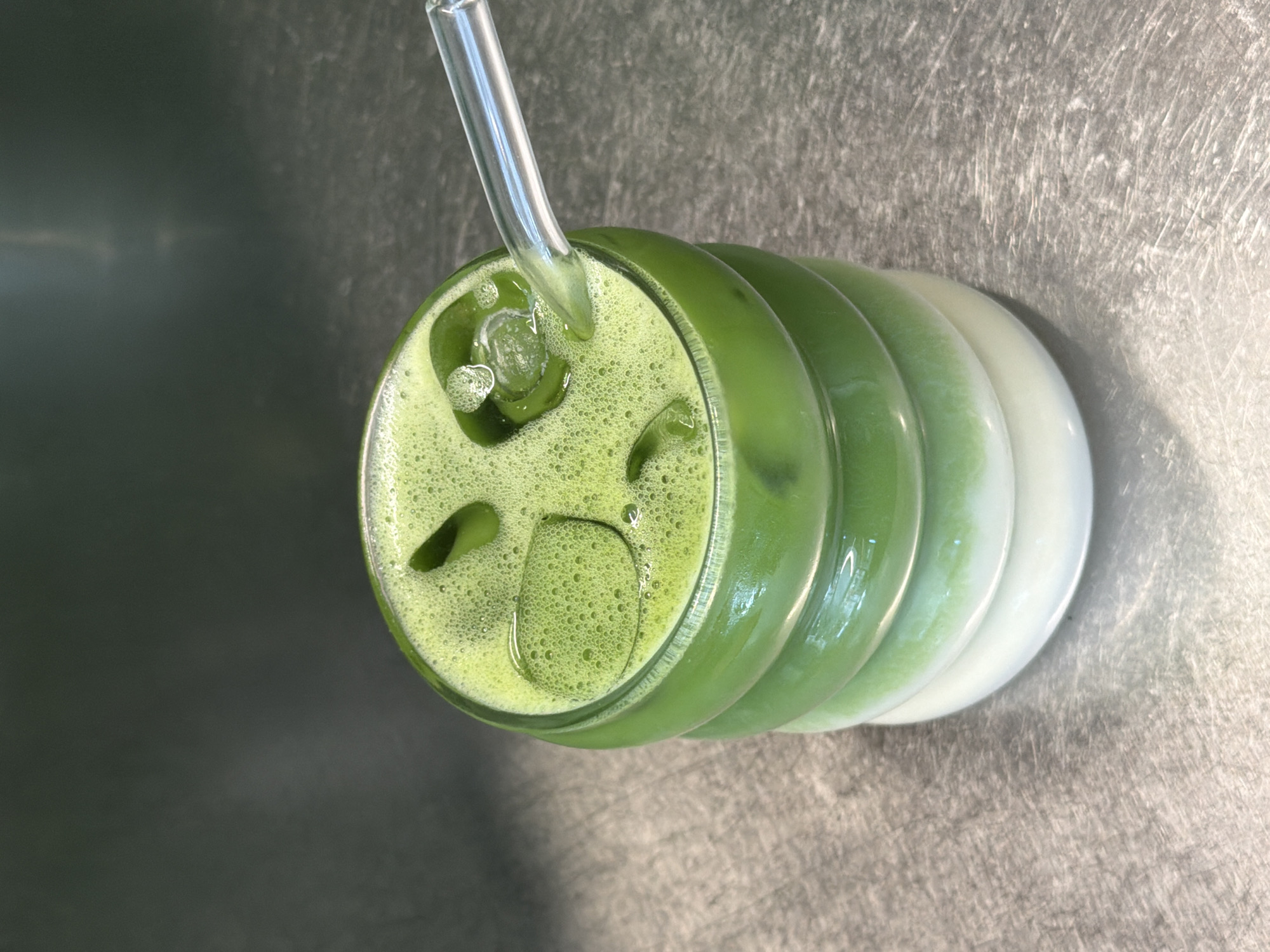 A close-up shot shows a layered green and white beverage in a clear, ribbed glass. The beverage is topped with ice cubes and foam, with a clear glass straw inserted into the top layer. The drink sits on a metallic surface, possibly a countertop. The background is dark and out of focus. The scene is set in an indoor environment, though the exact location within Sofia, Bulgaria is not discernible from the image. There are no people or animals present.