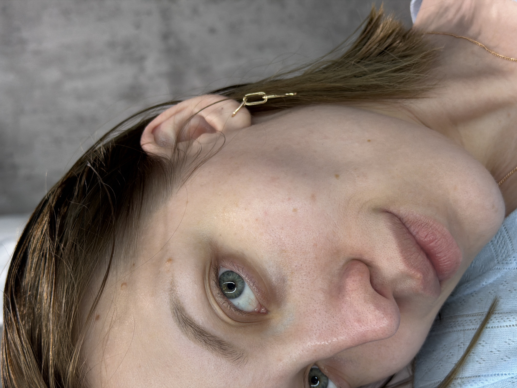 A close-up, tilted, full-frame view captures a woman's face, with focus primarily on her left side. Her eyes are green, and her skin exhibits freckles. She is wearing a gold-colored hair clip shaped like a chain link at the top of her ear. She also wears a thin gold chain around her neck. Her light-brown hair is visible, and she appears to be lying down on a light blue fabric surface. The background consists of a blurred gray surface. The location or city in Russia is not discernible from the provided image.