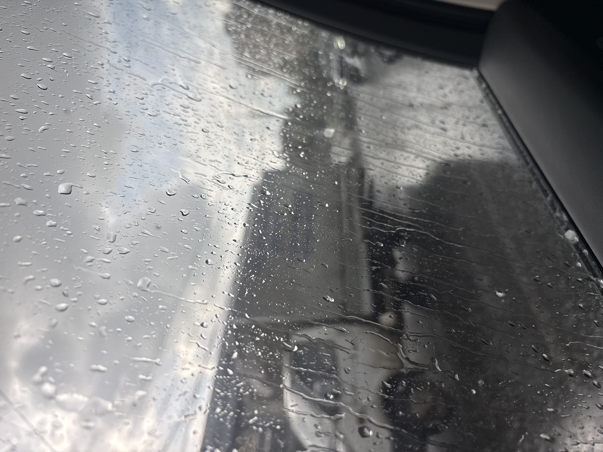 View through a raindrop-covered car window. Outside, there is a blurry cityscape, possibly in Yerevan, Armenia, although specific landmarks are obscured. The sky is overcast. The image contains no discernible people or actions. The environment is wet due to rain.