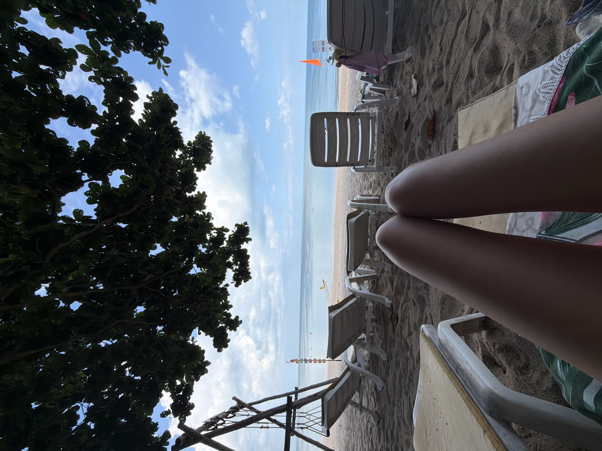 A first-person perspective shot captures a serene beach scene in the Ko Samui District, Thailand. The foreground is dominated by a person's legs, resting on a sandy surface, with a beach towel draped over them. In the middle ground, several empty lounge chairs are arranged on the sand, facing the ocean. A swing set stands to the left, adding a touch of playful ambiance. The background features a calm sea under a bright blue sky dotted with scattered clouds. The overall impression is one of relaxation and tranquility.