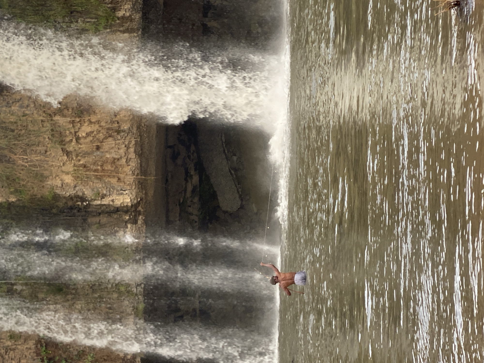 The image shows a waterfall scene. A man stands in the water near the base of the falls with both arms raised to different heights. The water is murky and reflects light. The waterfall flows from a rocky cliff face. The image appears to be rotated. Kukum, Nigeria is not discernible from the image content.