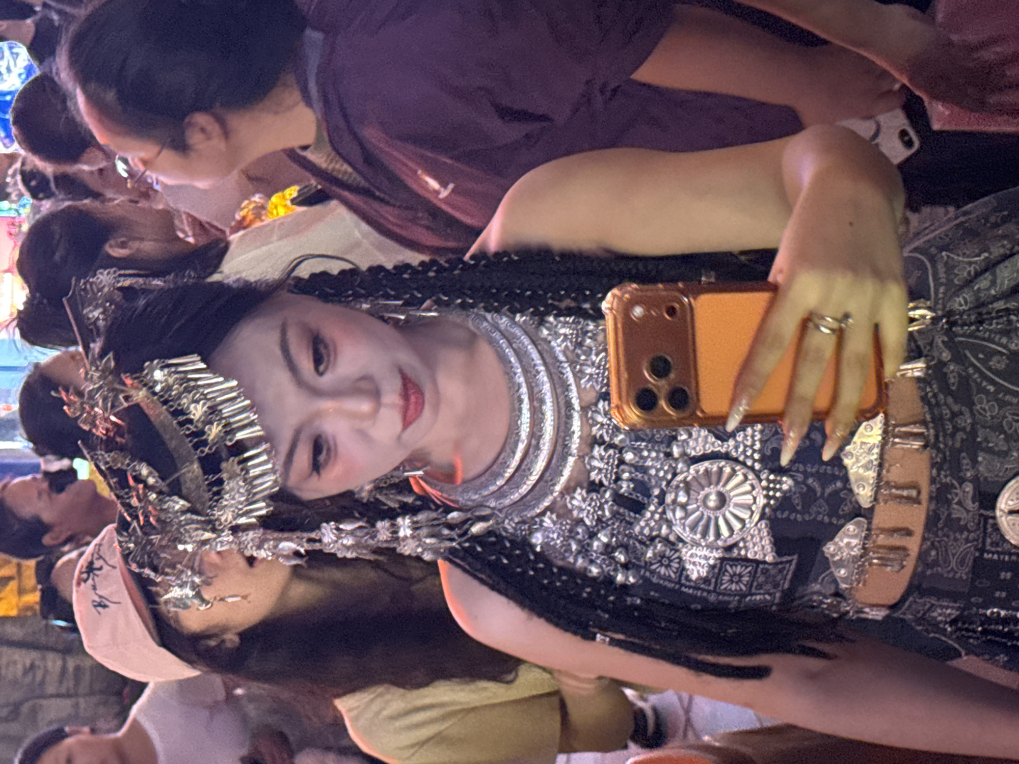 In Yuzhong District, China, a young woman dressed in traditional attire and elaborate silver jewelry is taking a selfie with her smartphone. She has dark braided hair adorned with a jeweled headpiece, heavy earrings, and a layered necklace. She is wearing a dark patterned top and a decorated belt. Behind her, other people are visible in the background, suggesting a public gathering or event. The lighting indicates it is likely nighttime or indoors with artificial lighting.