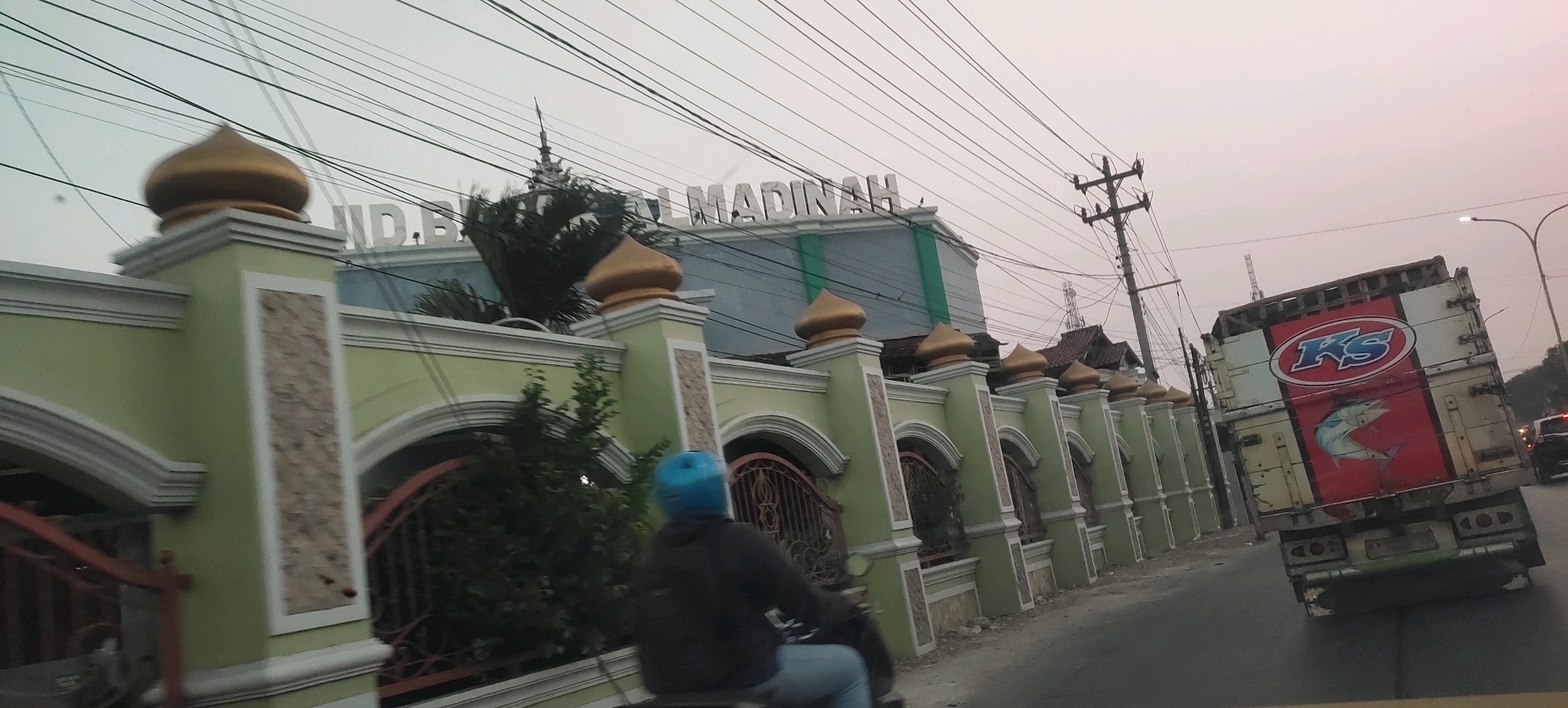 The image captures a street scene in Karanganyar, Indonesia, with a focus on the building facade and a passing truck. A light green fence with decorative arches and golden domed finials lines the street, separating it from a building. The building itself has a modern, multi-story design with large windows and a prominent sign reading "MADINAH" in white lettering. Electrical wires crisscross the sky above. In the foreground, a person wearing a blue helmet rides a motorcycle, moving away from the viewer. To the right of the fence, a large truck with a red and white design featuring a fish and the letters "KS" is driving on the road. The sky is overcast with a hazy, pale light.