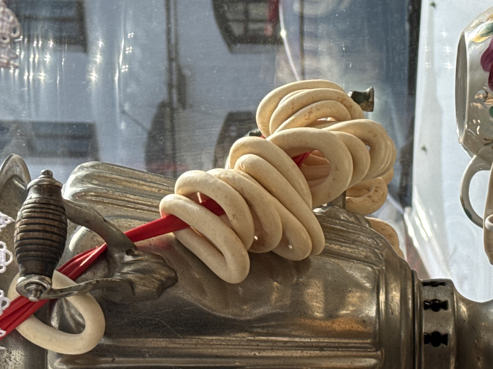 A close-up shot depicts a bunch of ring-shaped beige biscuits threaded on a red ribbon, likely displayed on a vintage metal meat grinder. The background is a blurred, possibly urban setting, perhaps observed through a glass display. The biscuits, typical of Russian cuisine, suggest a location where such food items are commonplace. The presence of the antique meat grinder further hints towards a traditional or historical setting, which aligns with the potential location of Kladovitsy, Russia, known for its cultural heritage.