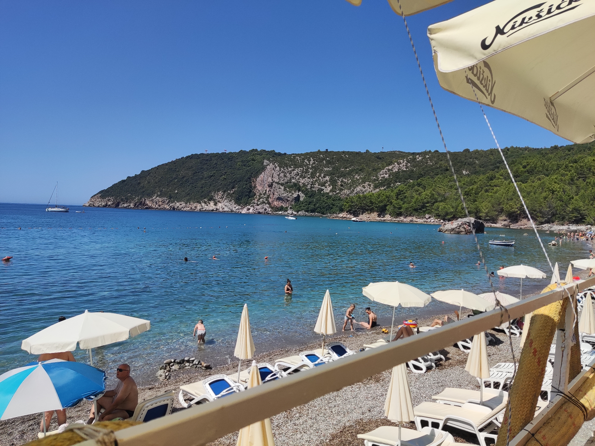 The image depicts a sunny day at a pebbled beach in Zagrađe, Montenegro, with clear blue water and a lush green, forested hillside in the background. The foreground is dominated by a wooden railing and a partial view of a light-colored awning with text. Several beach umbrellas, mostly white and light beige, are scattered across the beach, some providing shade for lounge chairs. People are visible on the beach and in the shallow water. Some are sunbathing on lounge chairs, while others are wading or swimming. A few individuals are interacting near the water's edge, and a child is standing in the shallow sea. In the distance, a sailboat is anchored in the bay, and a few small boats are also present. The overall scene is one of a leisurely summer day at the coast.