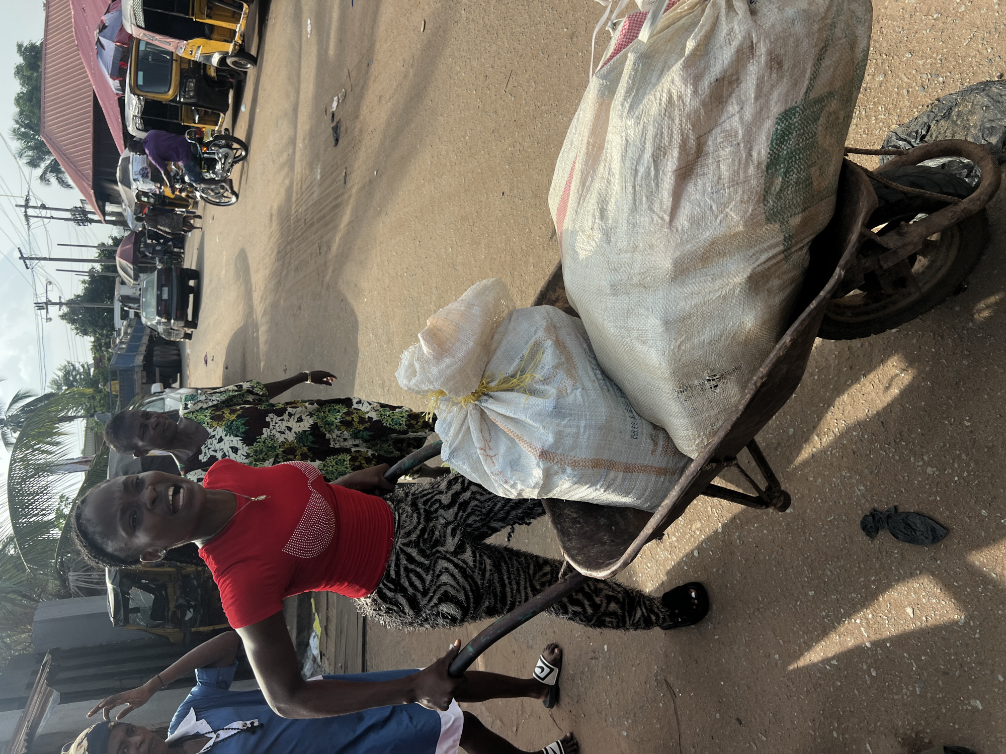 This image depicts a bustling street scene in Obosi, Nigeria, during bright daylight, likely late morning or early afternoon given the strong shadows. The central subjects are three individuals transporting goods. A young woman in a red t-shirt adorned with white embellishments and distinctive black and white patterned trousers is prominently featured, smiling broadly as she holds the handles of a heavily loaded, rusty metal wheelbarrow. Another person, partially visible in the foreground wearing a blue top, also assists in pushing the wheelbarrow.
The wheelbarrow carries two large, white woven sacks, one of which has a faint green design, possibly indicating "PRODUCE." Behind them, an older woman in a green, white, and black floral dress walks, looking towards the left. The background reveals a lively thoroughfare with several vehicles, including yellow tricycles (Keke Napep), motorcycles, and cars, suggesting a marketplace or active urban area. Buildings with reddish-brown roofs, utility poles, and tropical foliage like palm trees are visible under a clear sky. The ground is a paved, somewhat dusty surface. The young woman's cheerful expression stands out amidst the physical task of transporting goods.