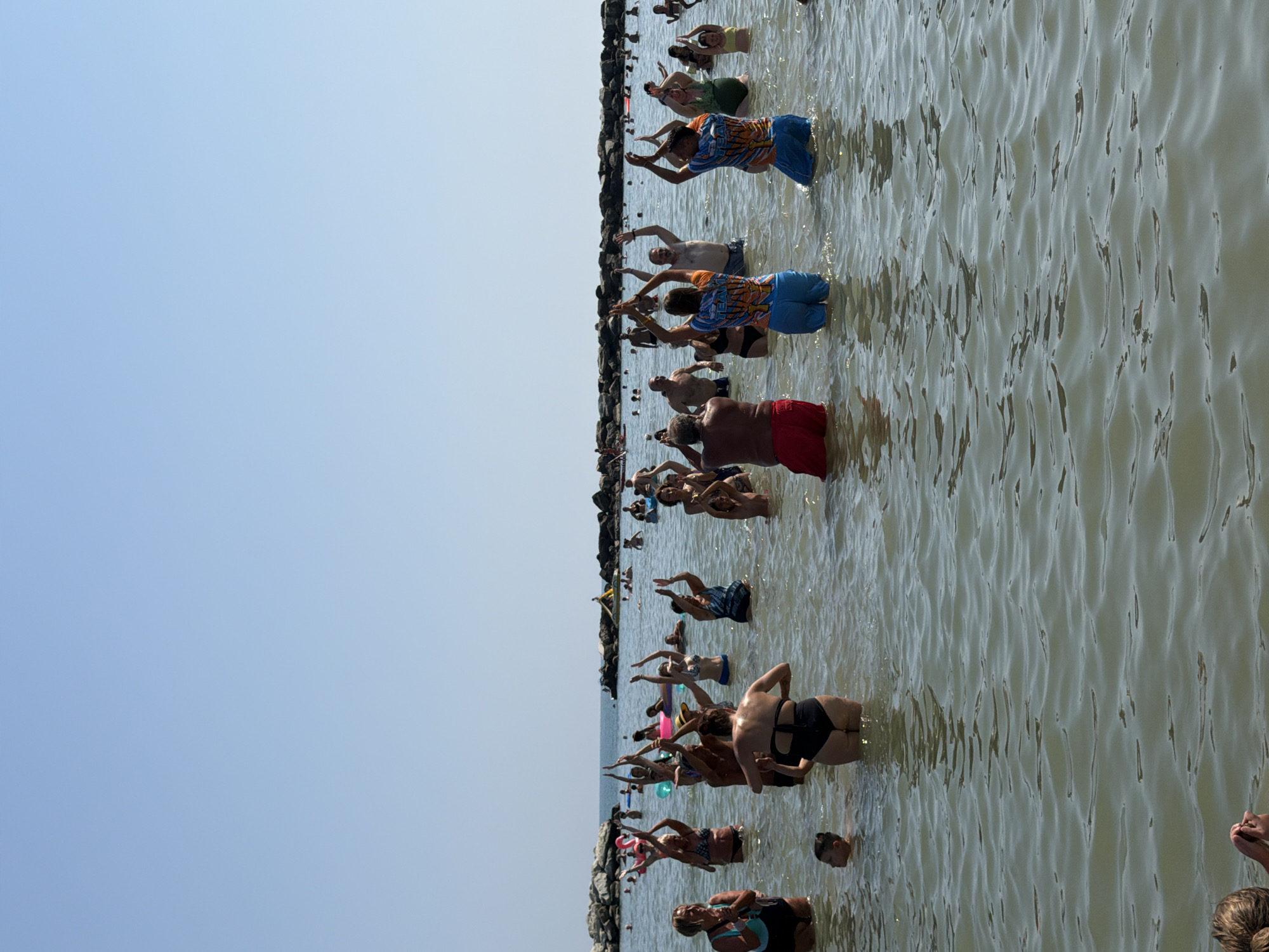 In the city of Gatteo, Italy, a group of people are standing in the shallow waters of the Adriatic Sea, possibly participating in a group exercise or yoga class. The sky is clear and blue, and the sun is shining. The people are of various ages and genders, and they are all wearing swimwear. Some are raising their hands in the air, while others are looking down or at each other. In the background, there are rock formations jutting out of the water, and a few more people can be seen further out in the sea. The water is calm, with gentle ripples on the surface.
