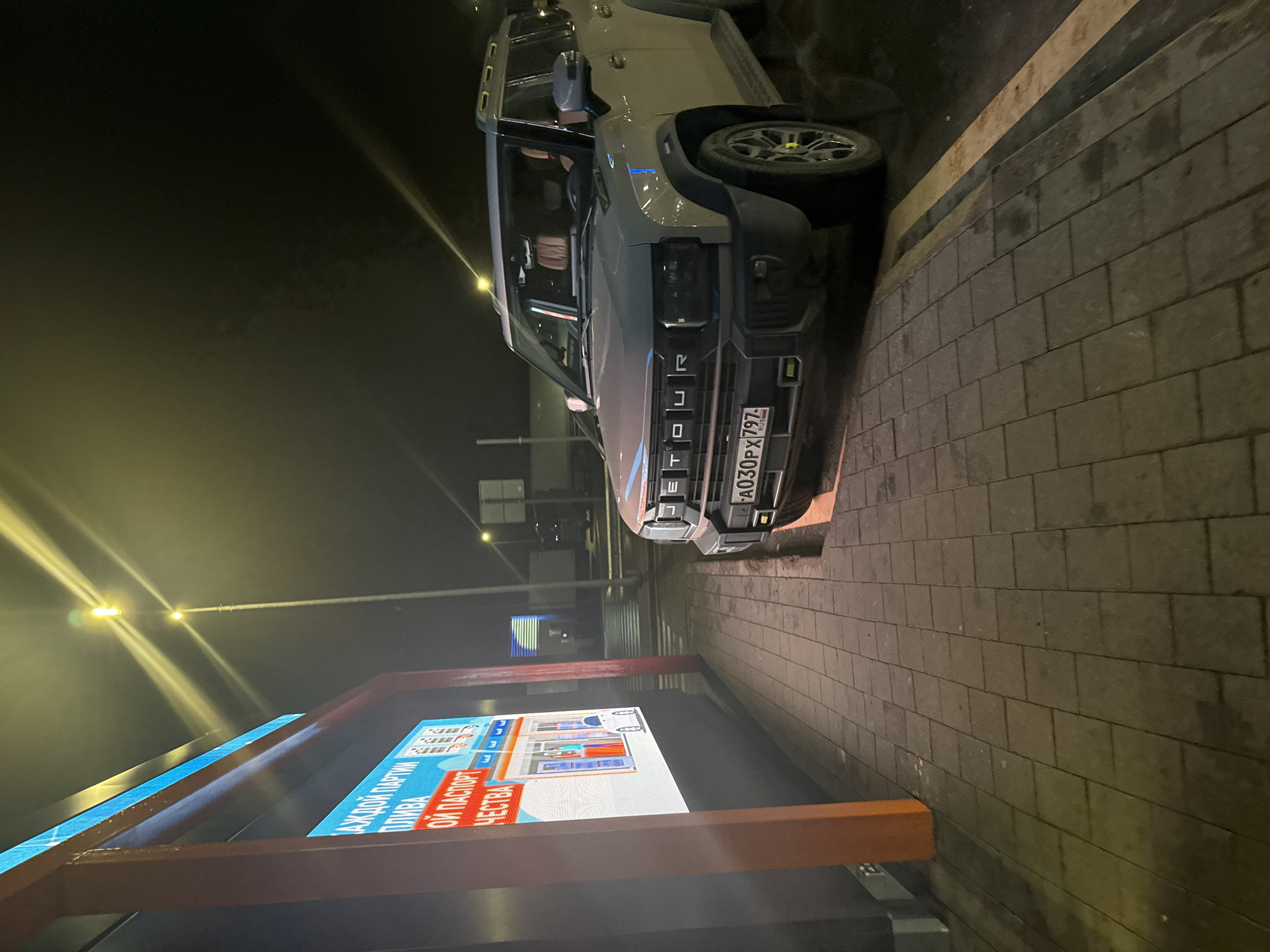 The image captures a scene in Bolshiye Dvory, Russia, at night. A light grey car, identified as a "TOUR" model by the lettering on its front grille, is parked on the side of a brick-paved road. The vehicle's license plate reads "A030PX 797 RUS," indicating its Russian registration. Next to the road, an illuminated advertising board displays text in Russian. Several streetlights are visible in the background, adding to the overall brightness of the scene.