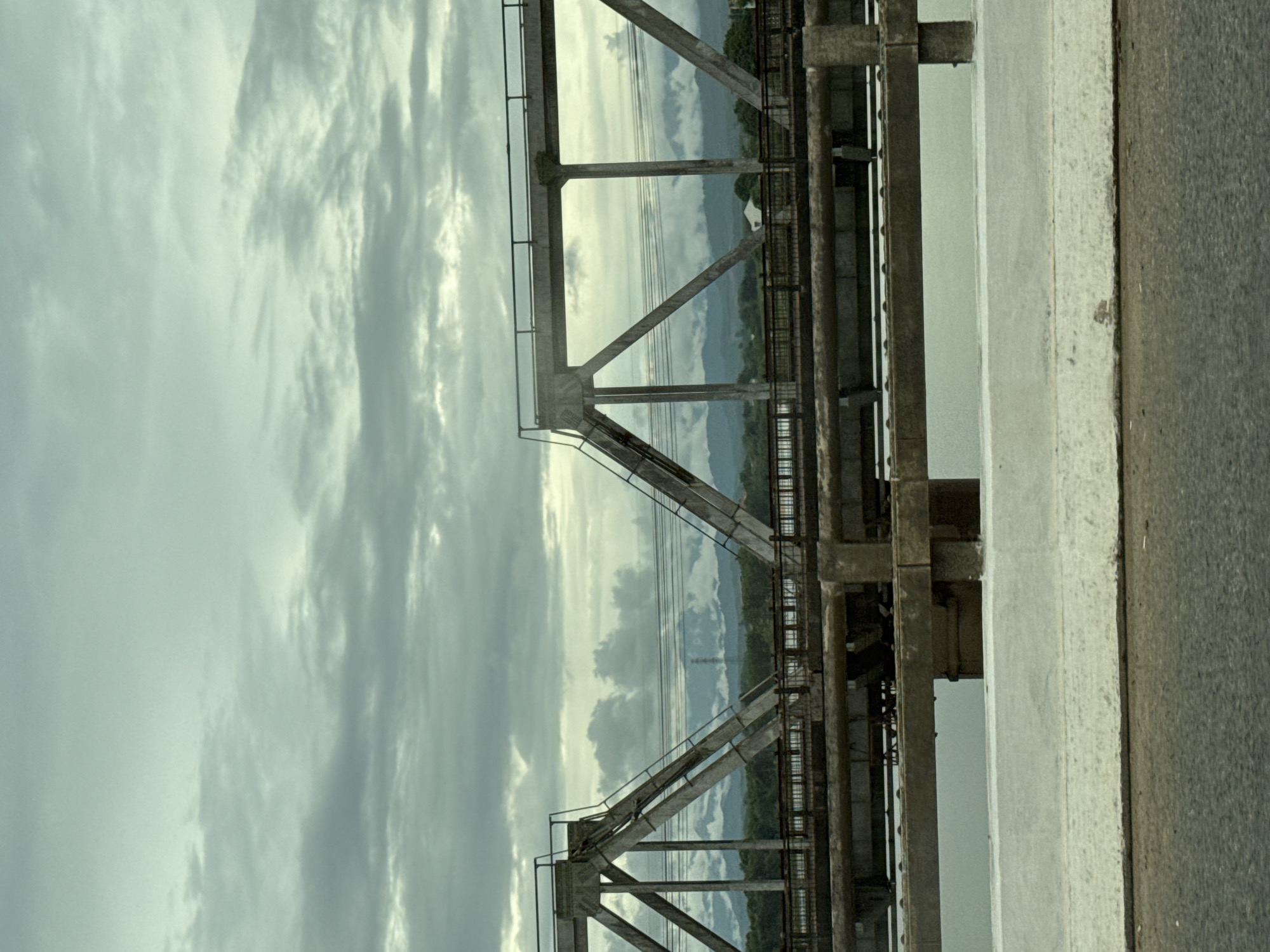 An overpass spans a body of water under a cloudy sky. The overpass is a truss bridge made of concrete and metal. The concrete pylons supporting the bridge are massive, and the metal beams are angled to form triangular supports. A lower deck of the bridge is visible beneath the main span, with a railing along its edge. Trees and distant mountains can be seen on the horizon beyond the bridge. The water beneath the bridge is calm and reflects the overcast sky. The foreground shows a section of a road, likely leading onto the overpass, with a concrete barrier separating it from the water. The overall impression is one of a functional, perhaps aging, piece of infrastructure in a natural setting. This scene is located in Quảng Trị Ward, Vietnam.